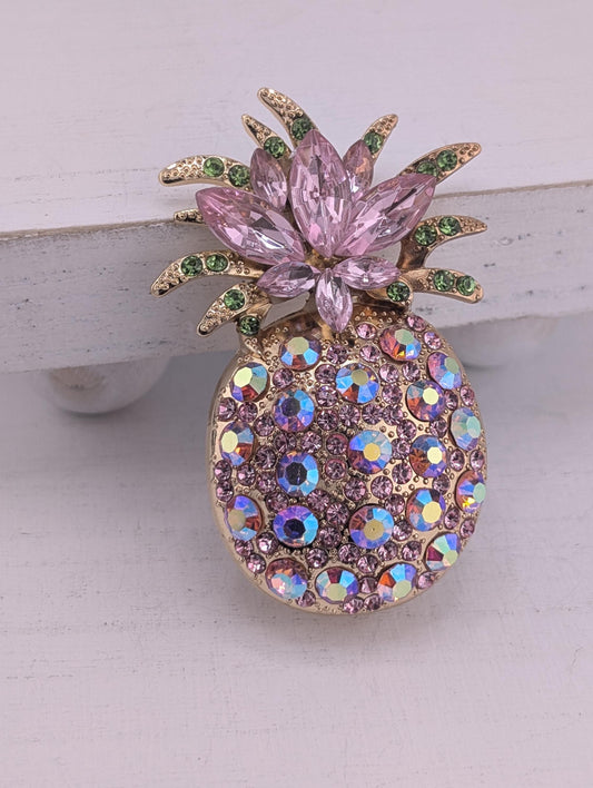 A decorative pin or brooch featuring a pineapple design with pink and purple gemstones, including a large flower-like element and smaller circular stones.