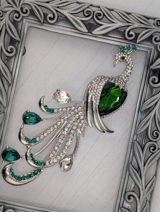 A decorative silver-framed brooch with a green gemstone and sparkling crystal accents.