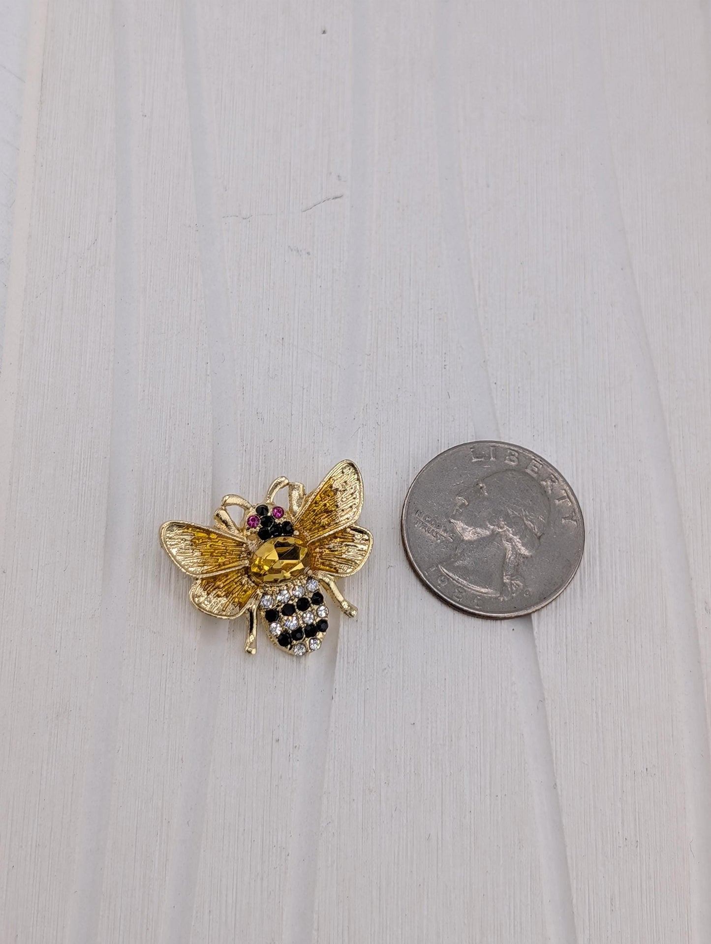 A gold-colored brooch in the shape of a bee is placed next to a silver coin on a white wooden surface.