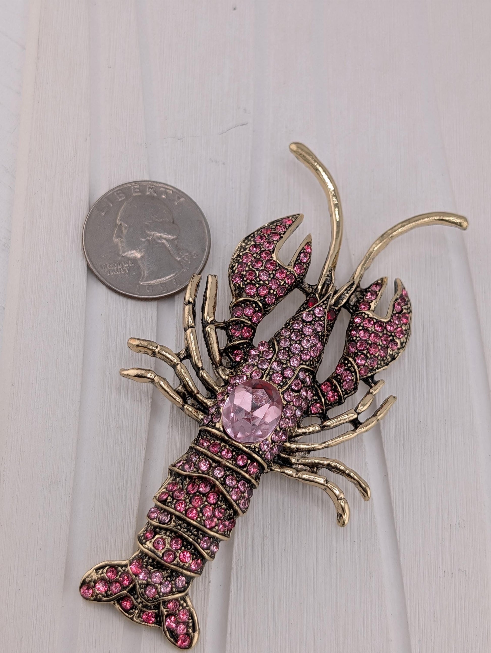 A pink rhinestone lobster brooch is placed next to a quarter on a white surface.
