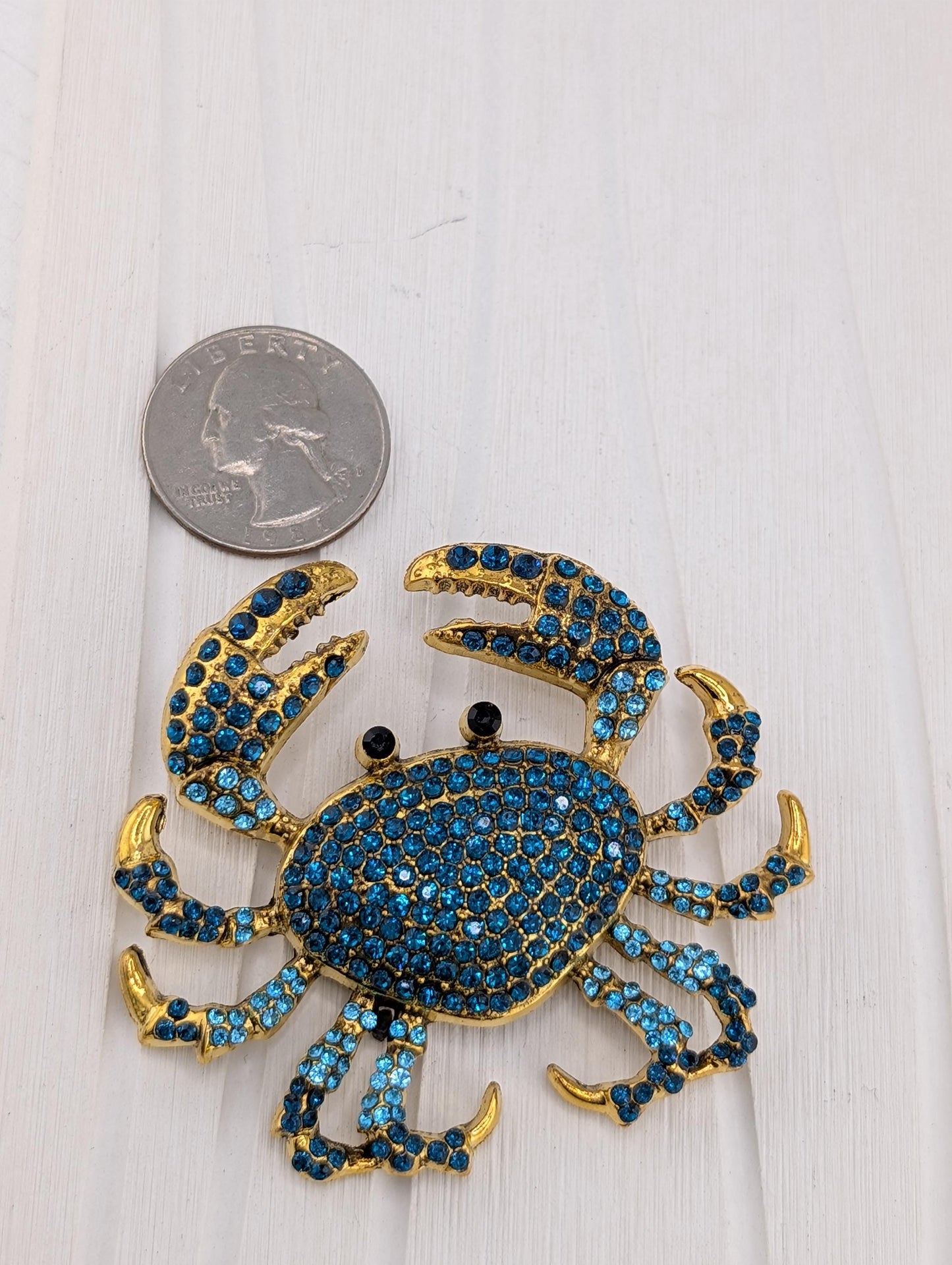 A shiny, blue crab-shaped brooch with gold accents and sparkling blue gemstones is placed next to a United States quarter on a white surface.