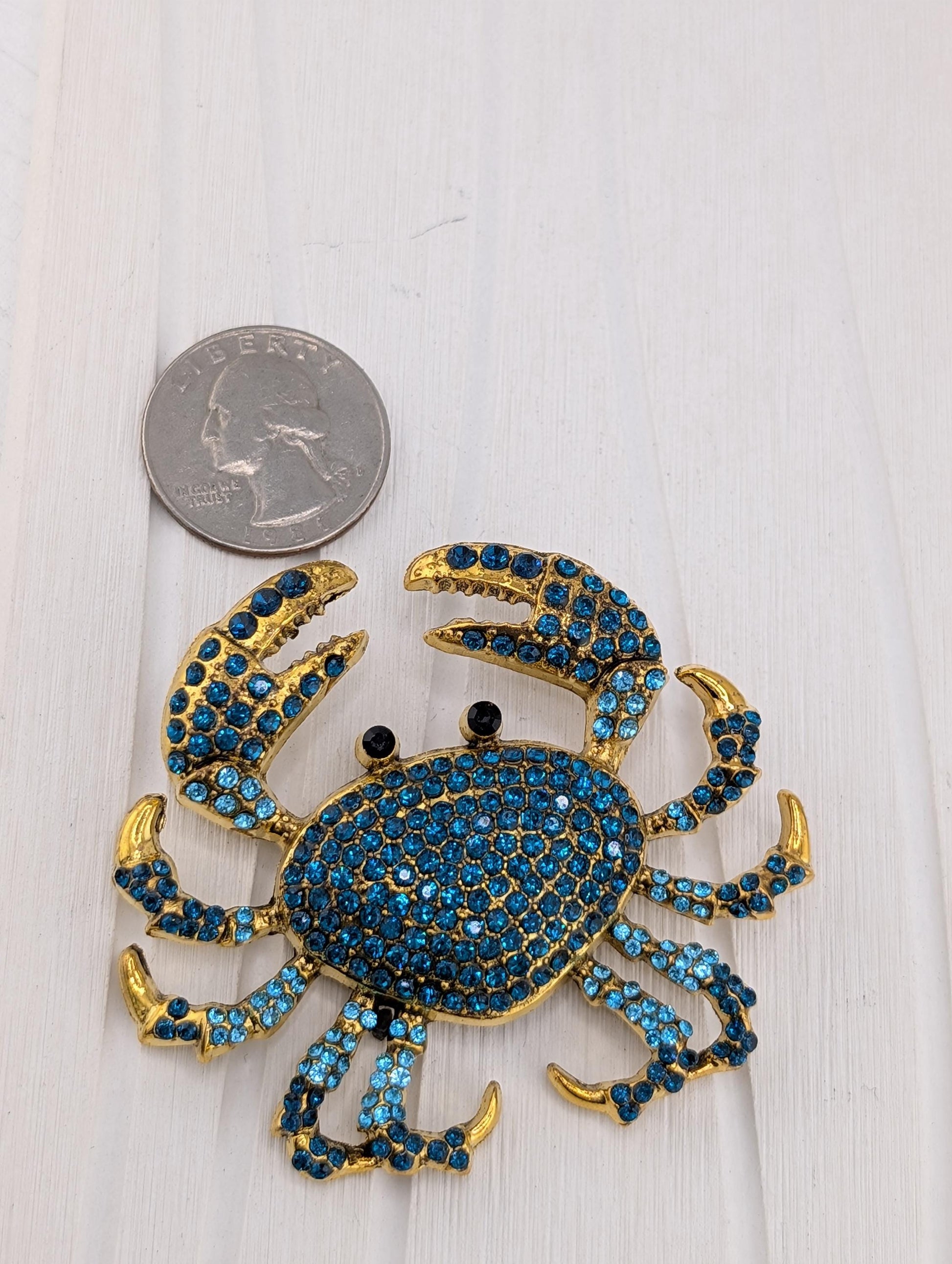 A shiny, blue crab-shaped brooch with gold accents and sparkling blue gemstones is placed next to a United States quarter on a white surface.