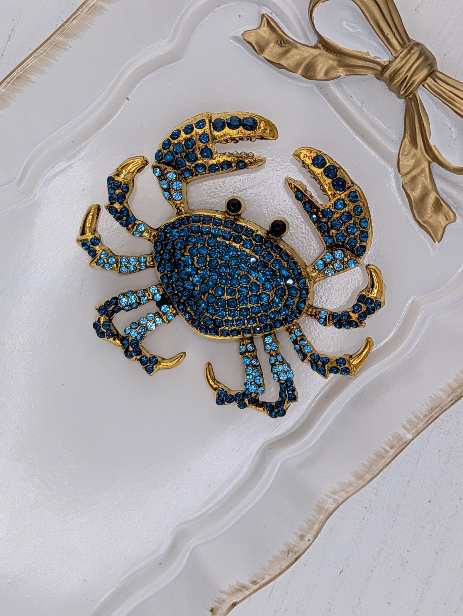 A decorative crab-shaped brooch with blue and gold gemstones is displayed on a white surface, accompanied by a gold ribbon bow.