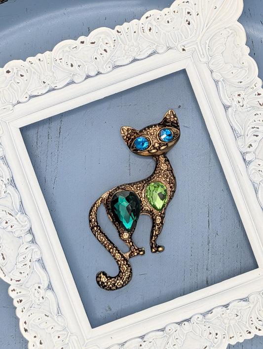 A decorative cat brooch with blue eyes and green gemstones is displayed in a white ornate frame against a blue background.