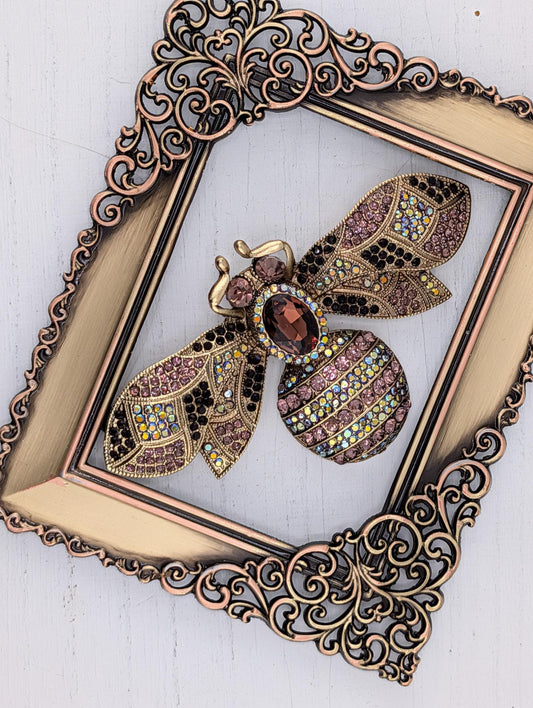 A decorative gold-framed picture of a colorful, ornate brooch in the shape of a bee.