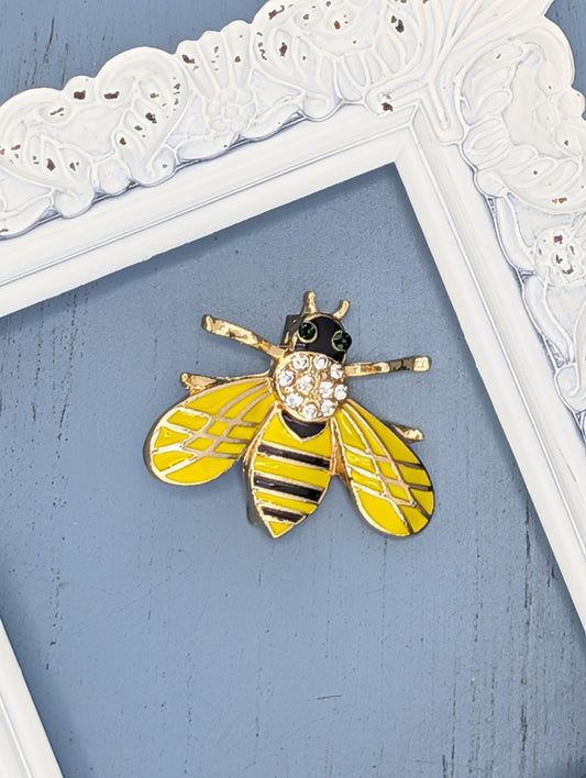 A decorative gold and yellow bee brooch is displayed on a blue background, framed by a white ornate frame.
