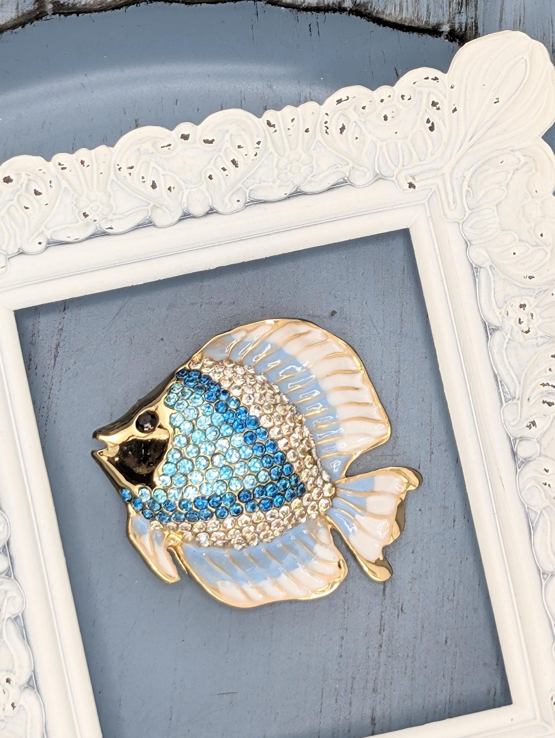 A decorative blue and gold fish brooch is displayed in a white ornate frame against a blue background.