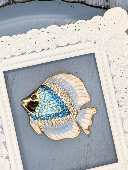 A decorative blue and gold fish brooch is displayed in a white ornate frame against a blue background.
