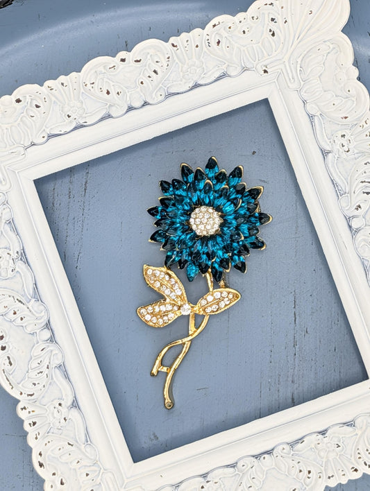 A decorative brooch in the shape of a blue flower with gold accents, displayed within a white ornate frame.