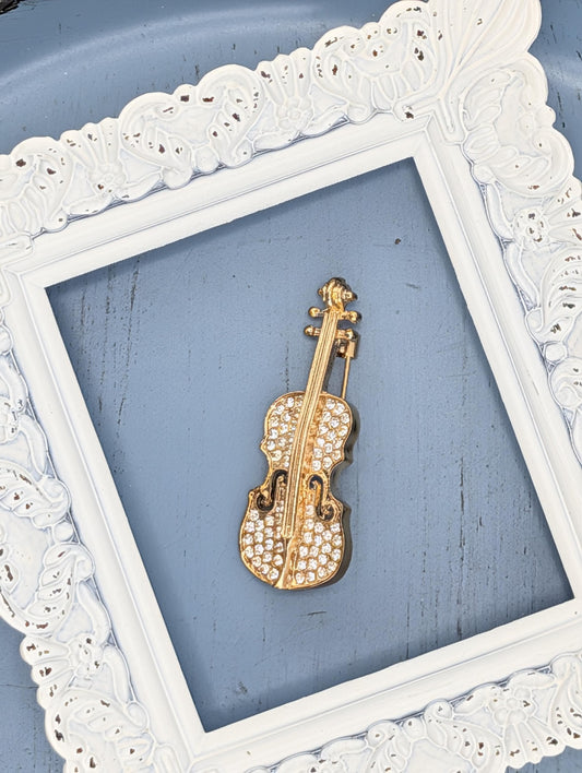 A gold-colored violin brooch with rhinestones is displayed in a white ornate frame against a blue background.