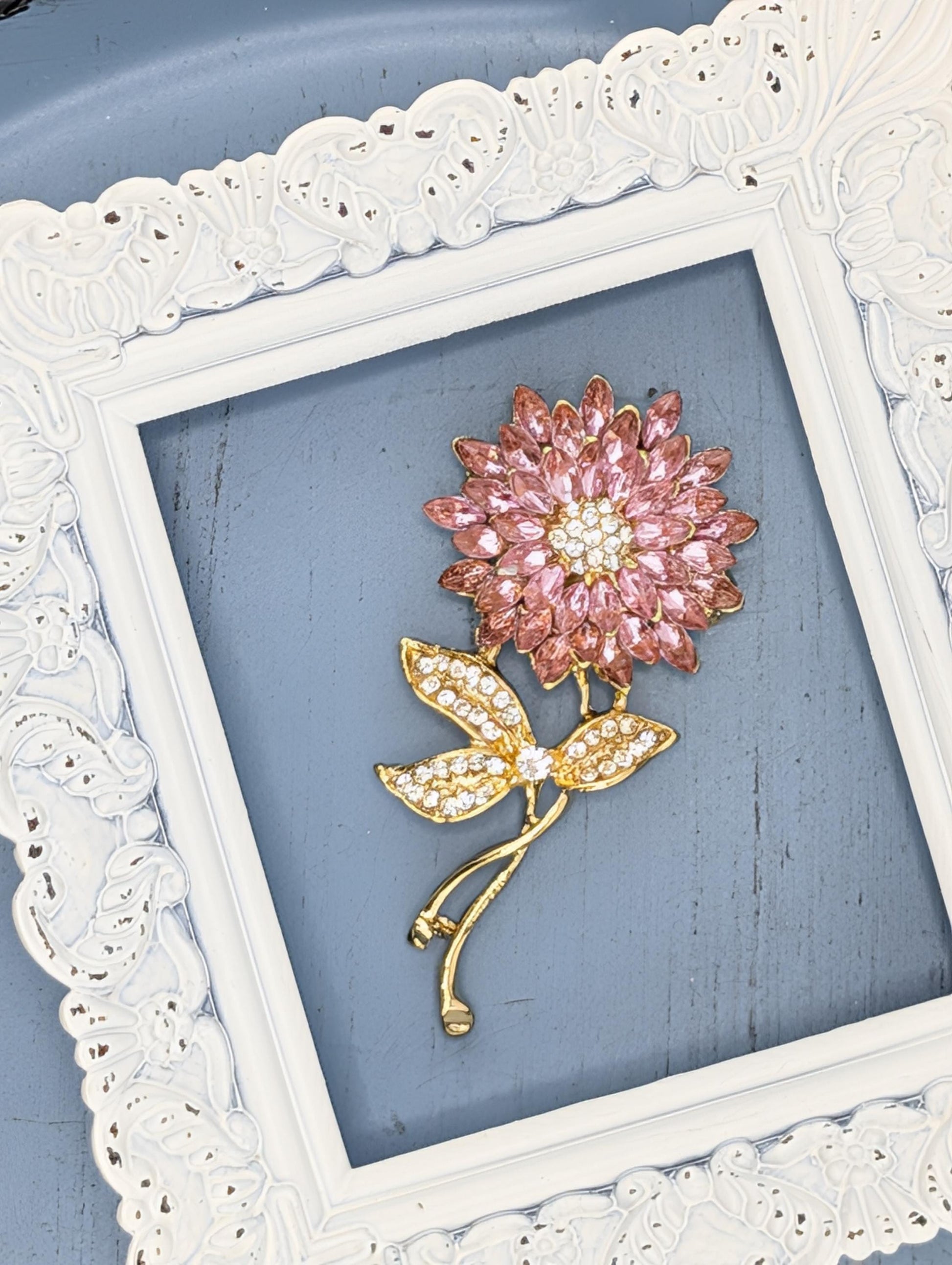 A decorative brooch in the shape of a flower, adorned with pink gemstones, is displayed within a white ornate frame against a blue background.