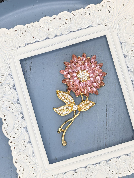 A decorative brooch in the shape of a flower, adorned with pink gemstones, is displayed within a white ornate frame against a blue background.