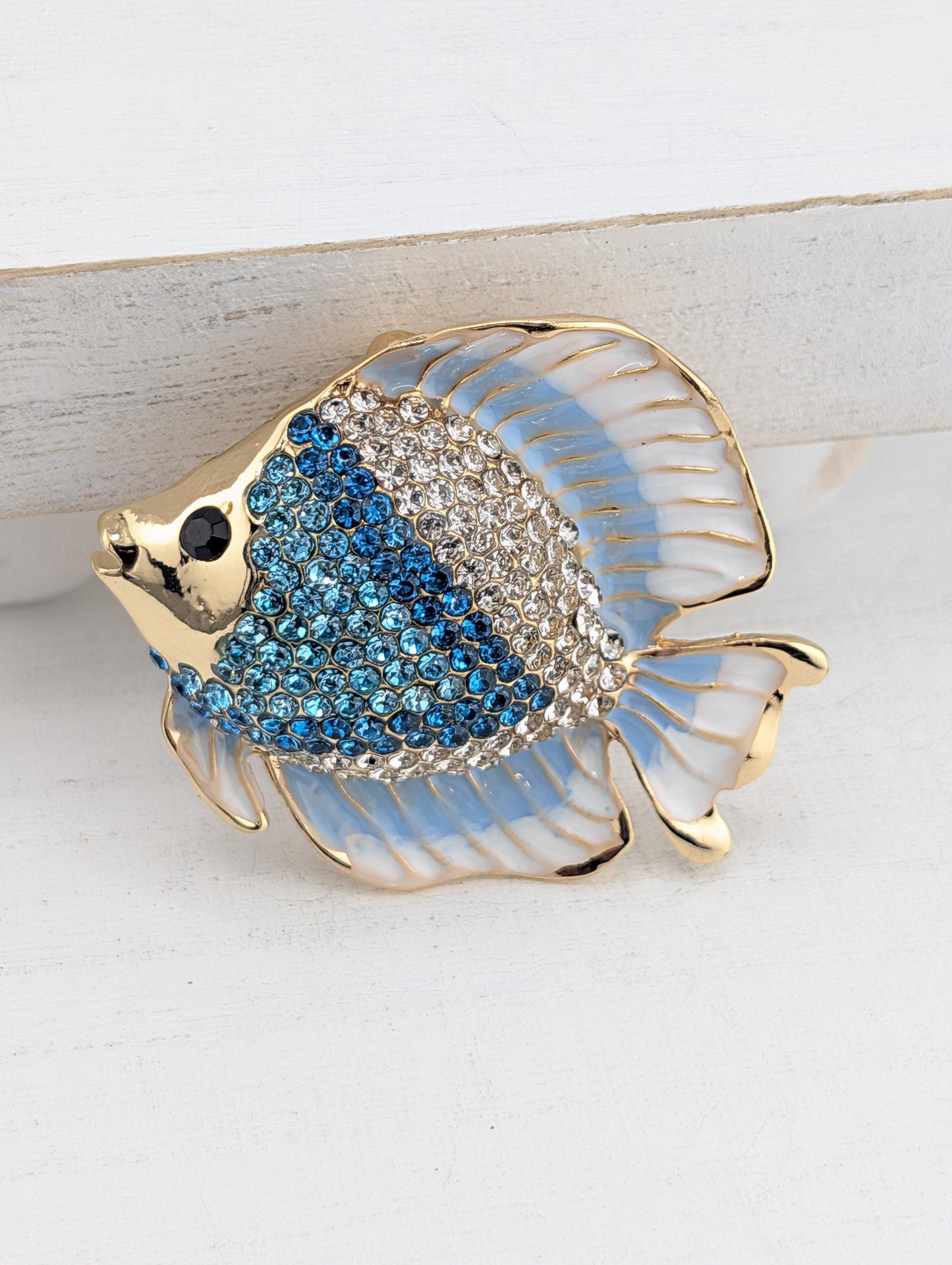 A decorative brooch in the shape of a fish, with blue and white colors and sparkling rhinestones or crystals on its body.