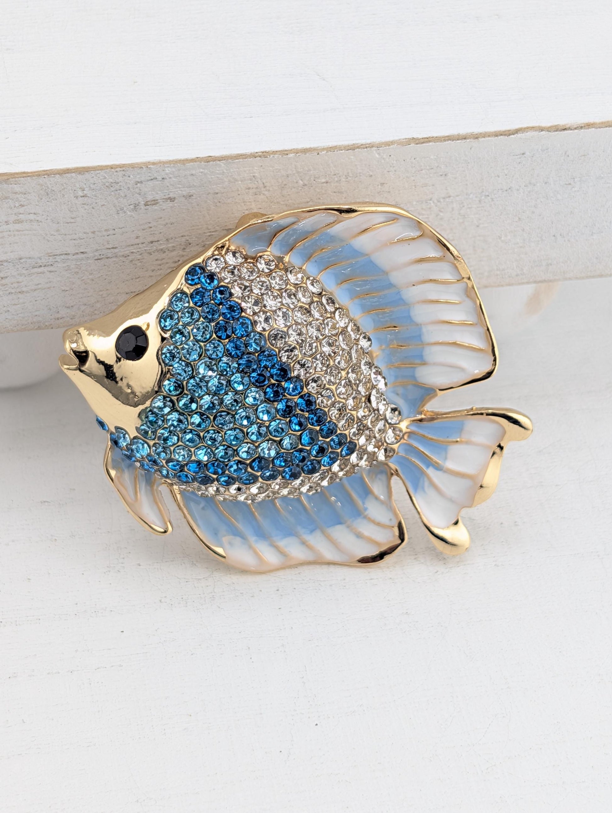 A decorative brooch in the shape of a fish, with blue and white colors and sparkling rhinestones or crystals on its body.