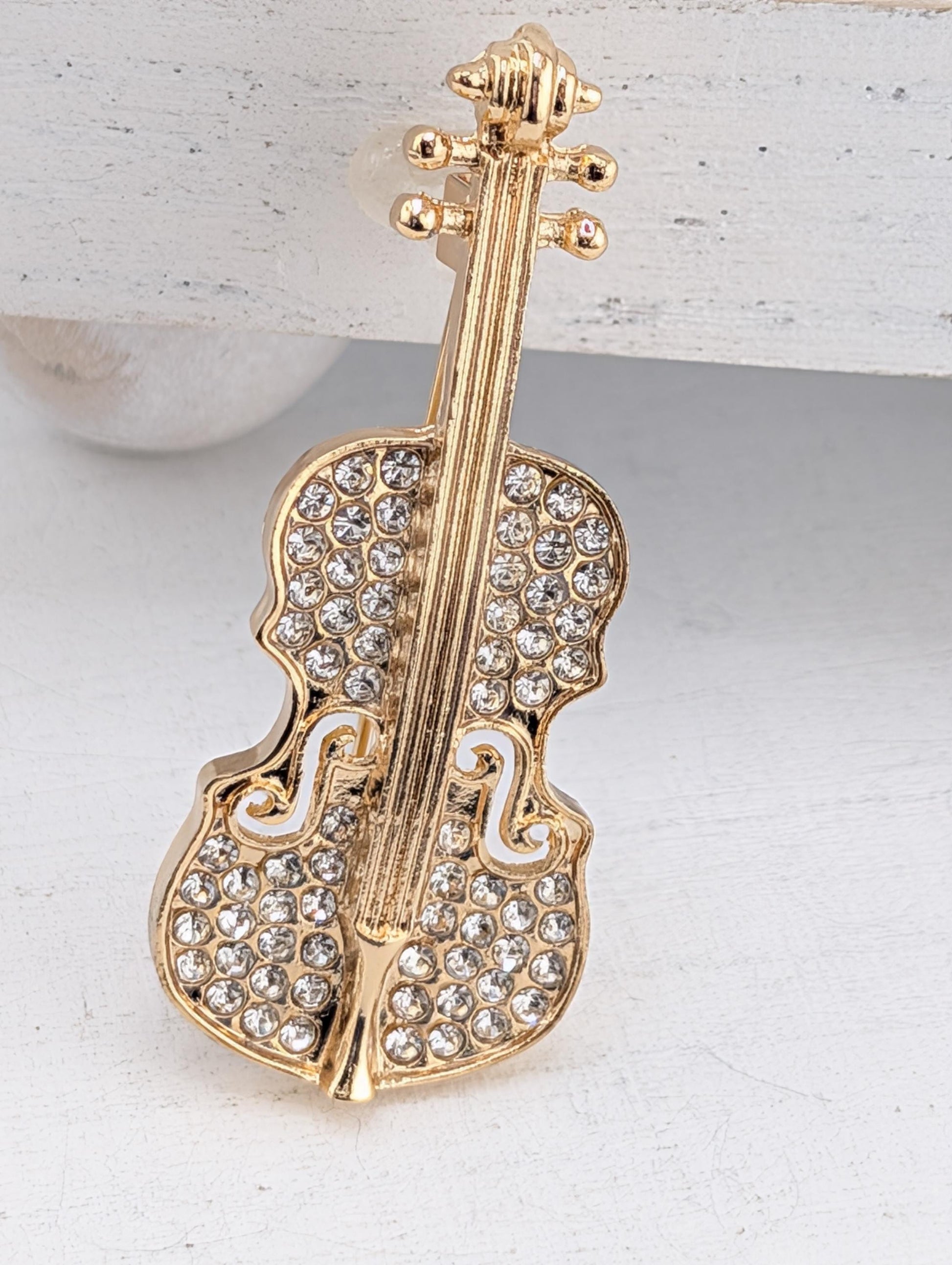 A gold-colored pendant in the shape of a violin, adorned with numerous small diamonds or crystals, is displayed against a white background.