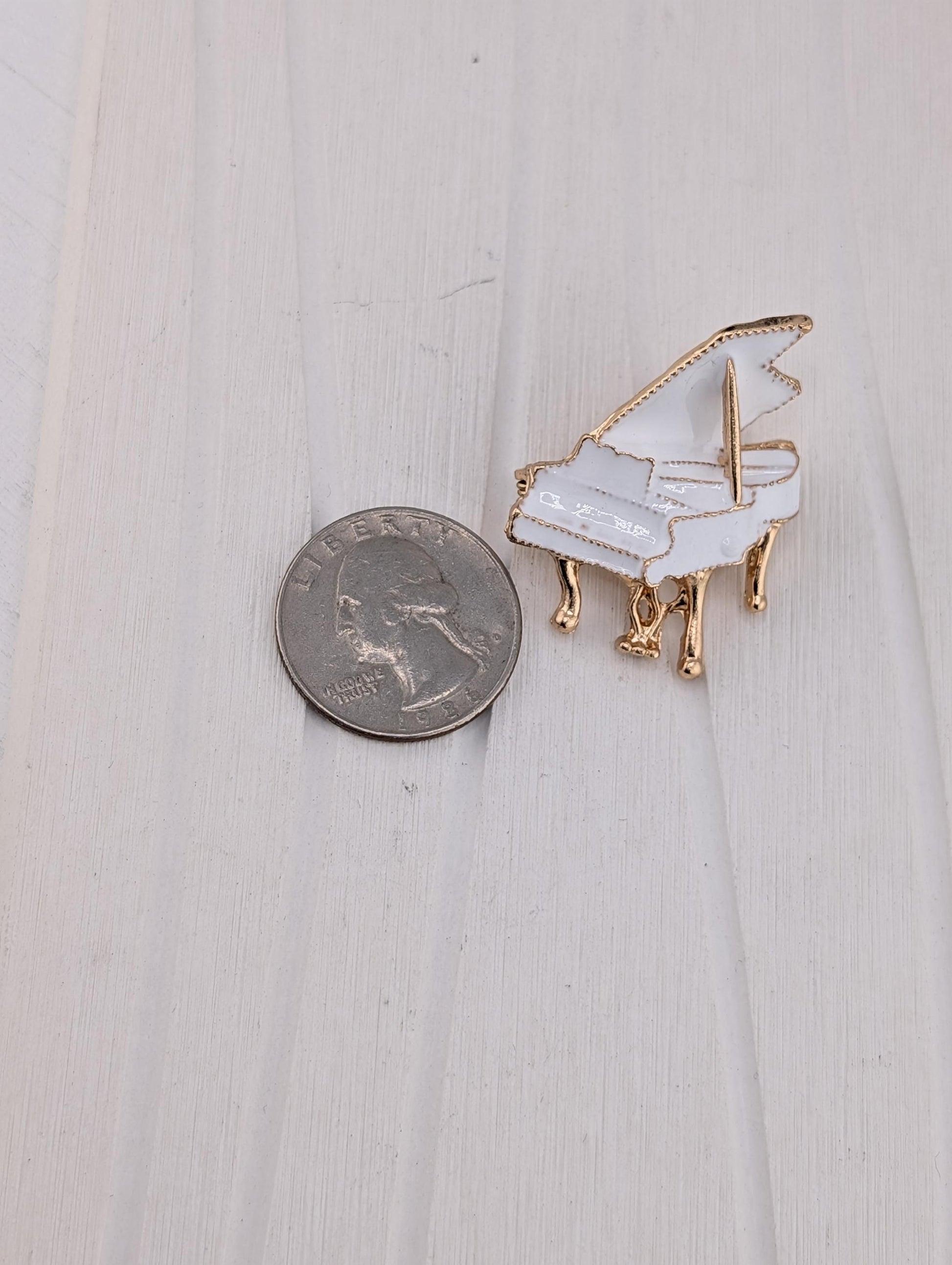 A small white and gold piano brooch is placed next to a United States quarter on a white wooden surface.