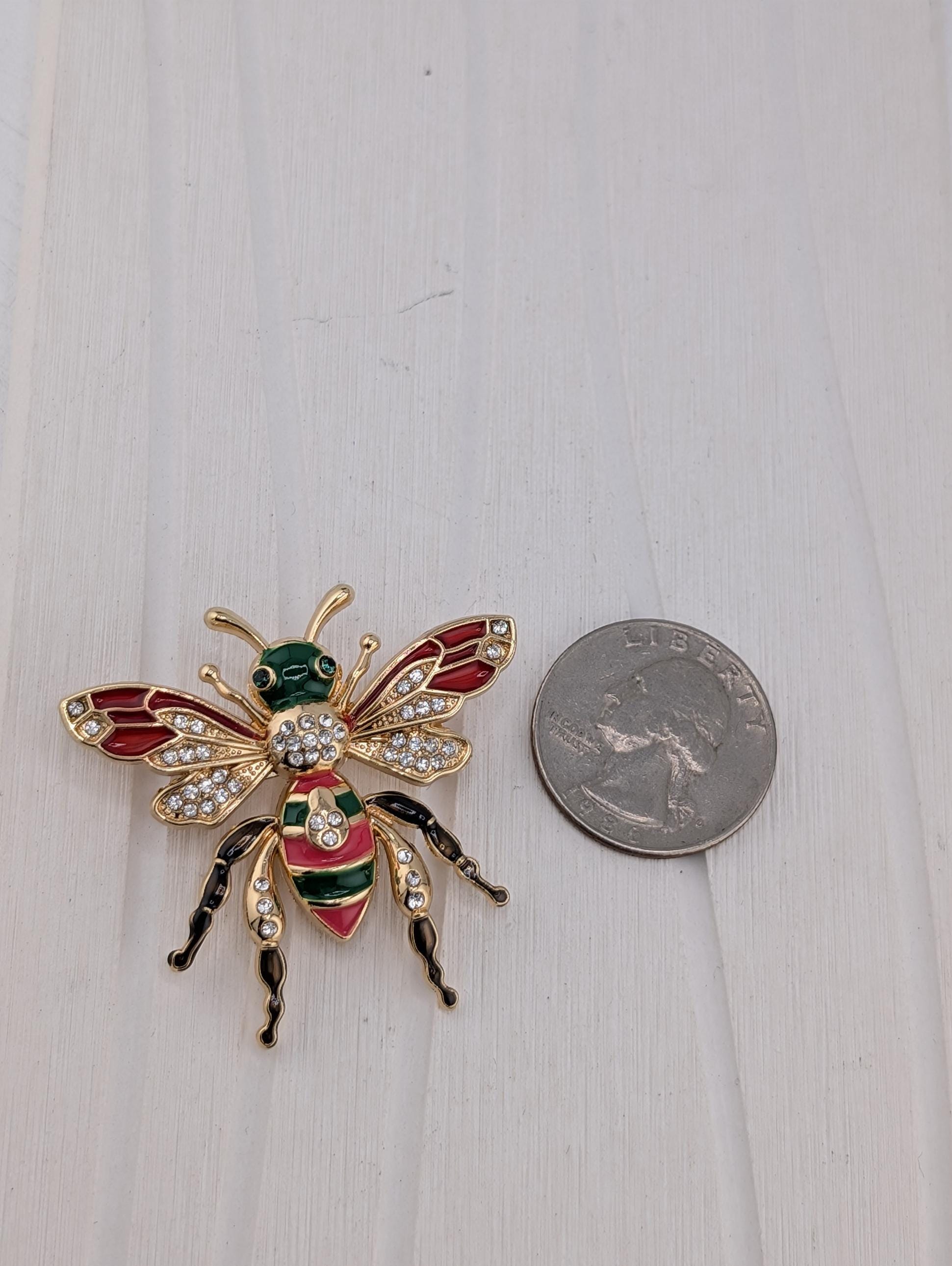 A decorative bee brooch with colorful wings and a green body is placed next to a US quarter on a white surface.