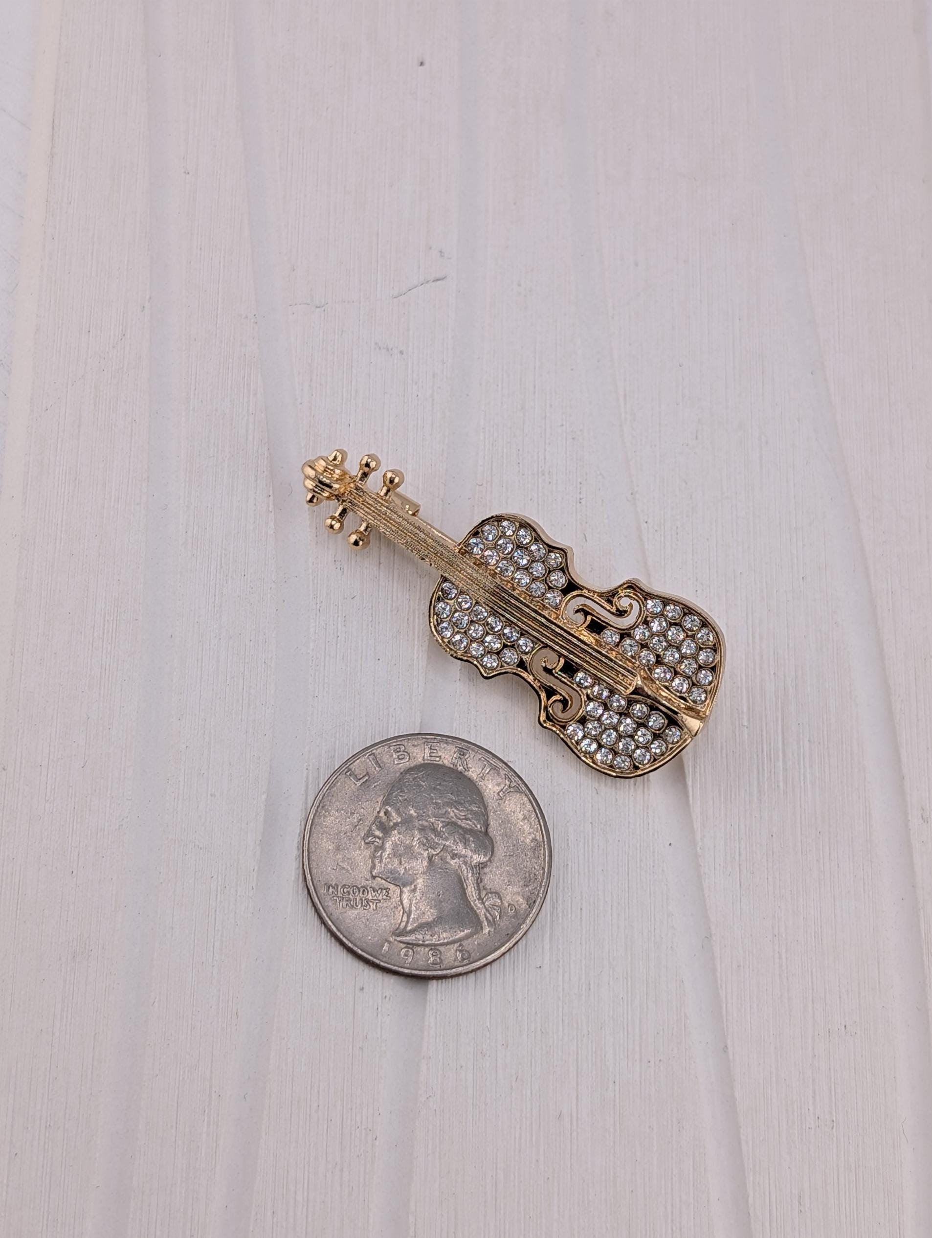 A gold-colored brooch with rhinestones is placed next to a silver-colored US quarter on a white wooden surface.