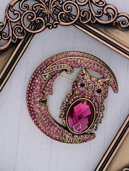 A pink gemstone owl brooch with a crescent moon design, set against a white background.