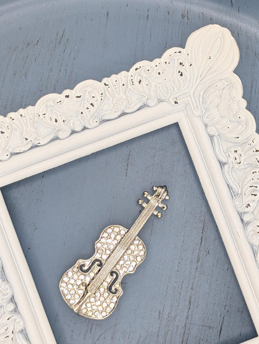 A white ornate frame with a decorative border is placed on a blue surface, with a small silver brooch in the shape of a violin resting on top of it.