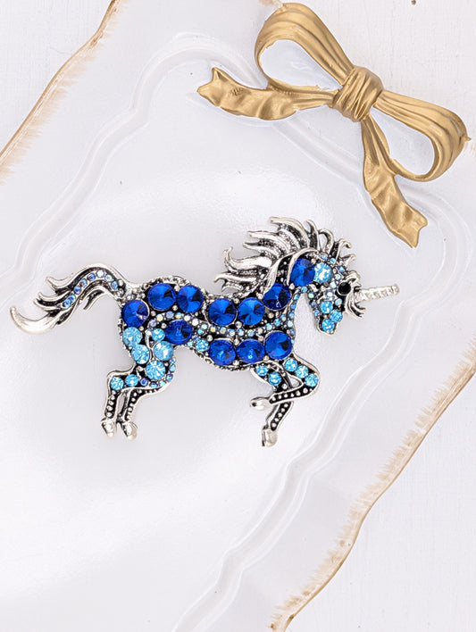 A decorative brooch in the shape of a unicorn, adorned with blue gemstones, is displayed against a white background.