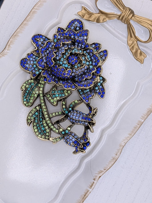 A decorative brooch with a large blue flower and green leaves, adorned with sparkling rhinestones.