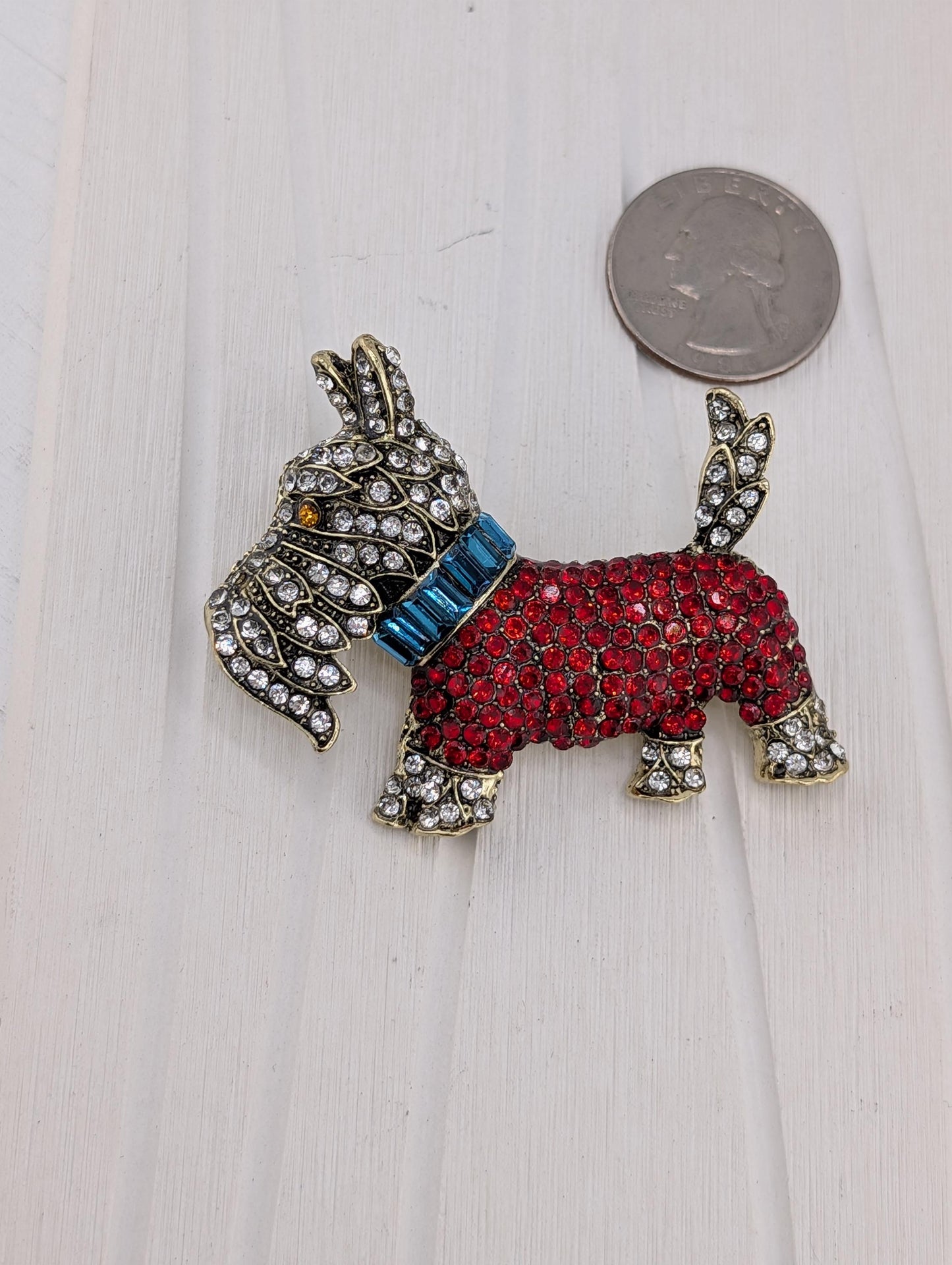 A decorative dog brooch made of red and blue gemstones, with a blue collar and a silver-colored coin placed next to it on a white surface.