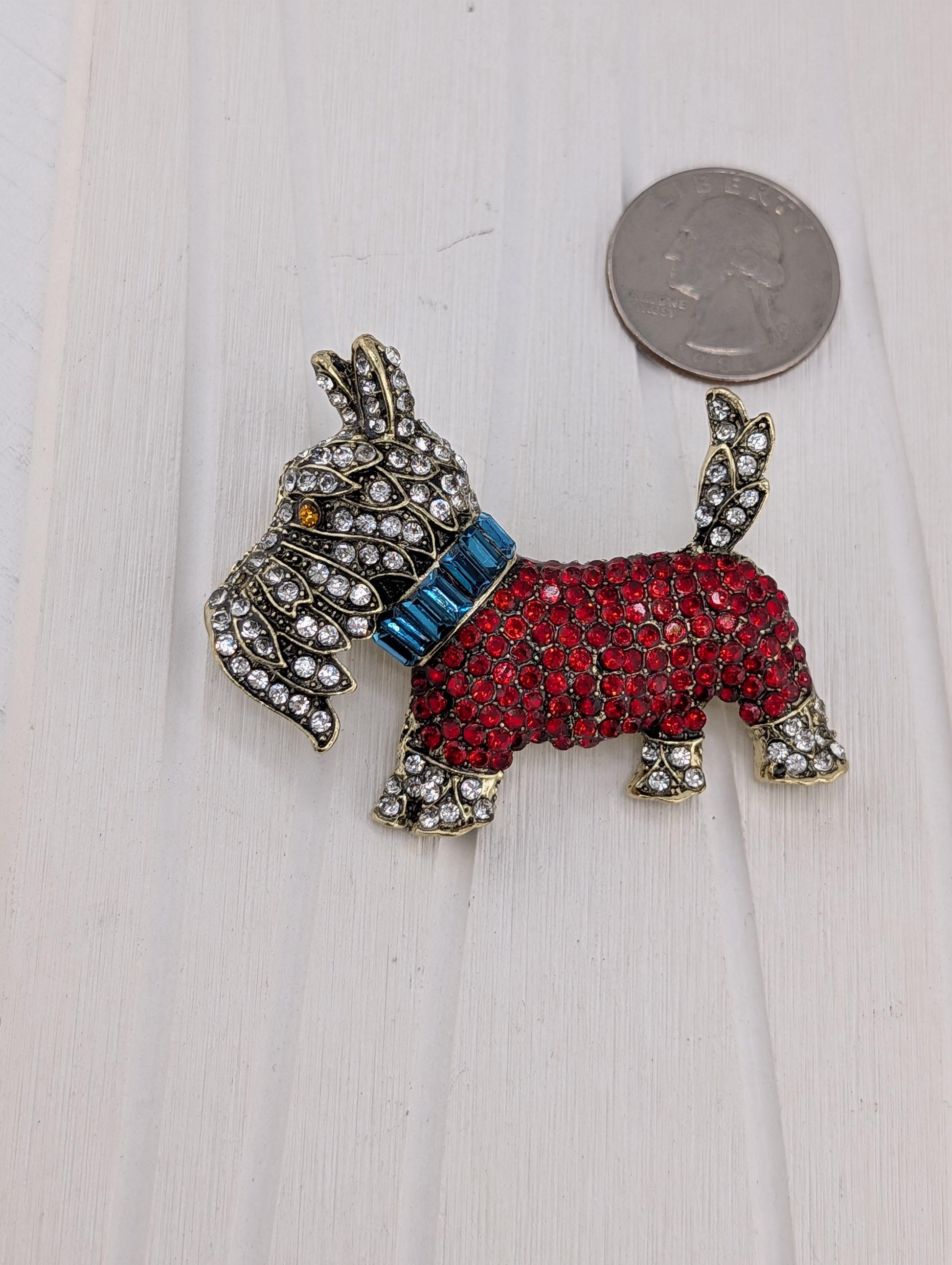 A decorative dog brooch made of red and blue gemstones, with a blue collar and a silver-colored coin placed next to it on a white surface.
