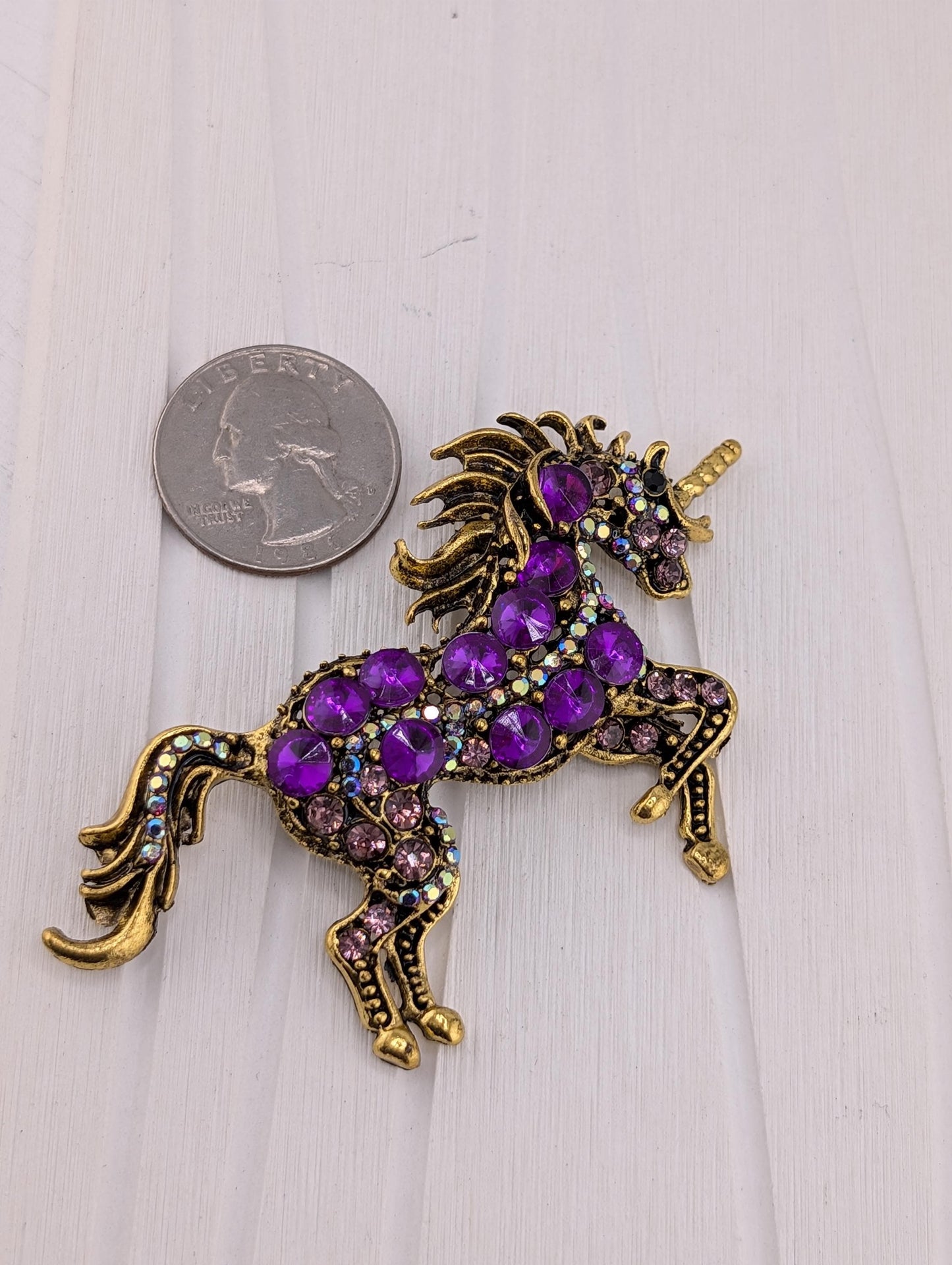 A decorative unicorn brooch adorned with purple gemstones and gold accents, placed next to a United States quarter coin.