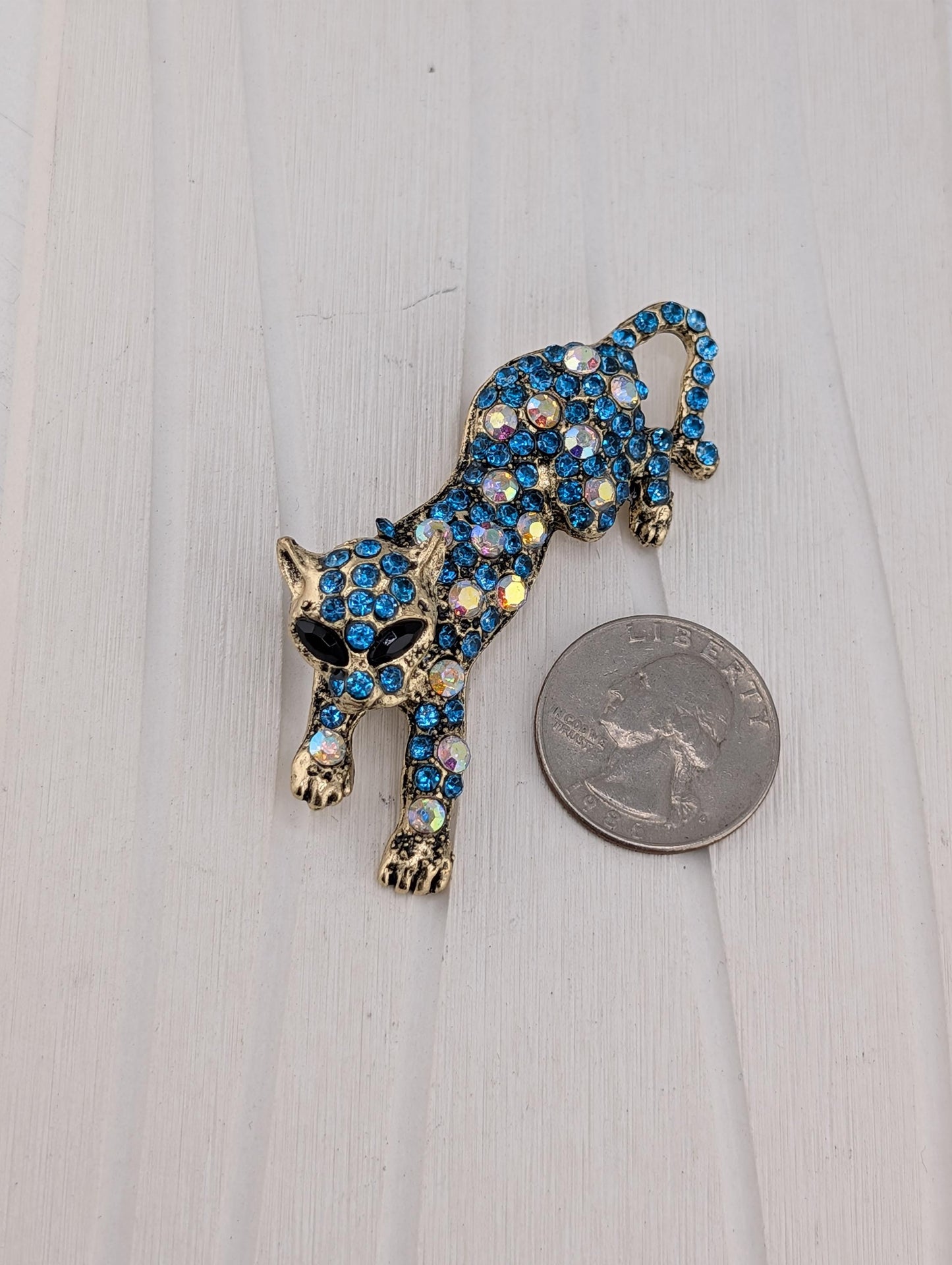 A decorative cat brooch made of blue and gold materials, adorned with sparkling rhinestones, is placed next to a United States quarter on a white surface.
