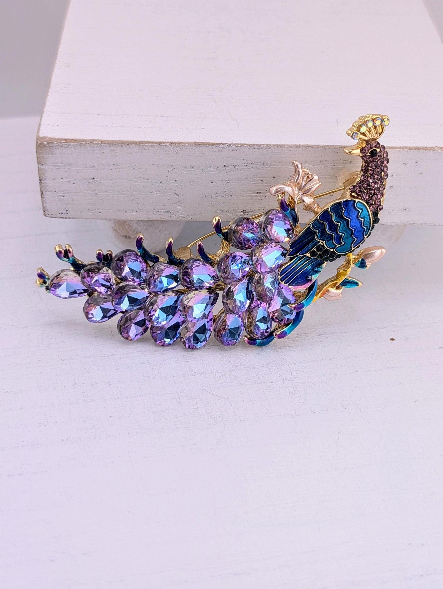 A decorative peacock brooch with a vibrant purple and blue color scheme, adorned with numerous sparkling gemstones.