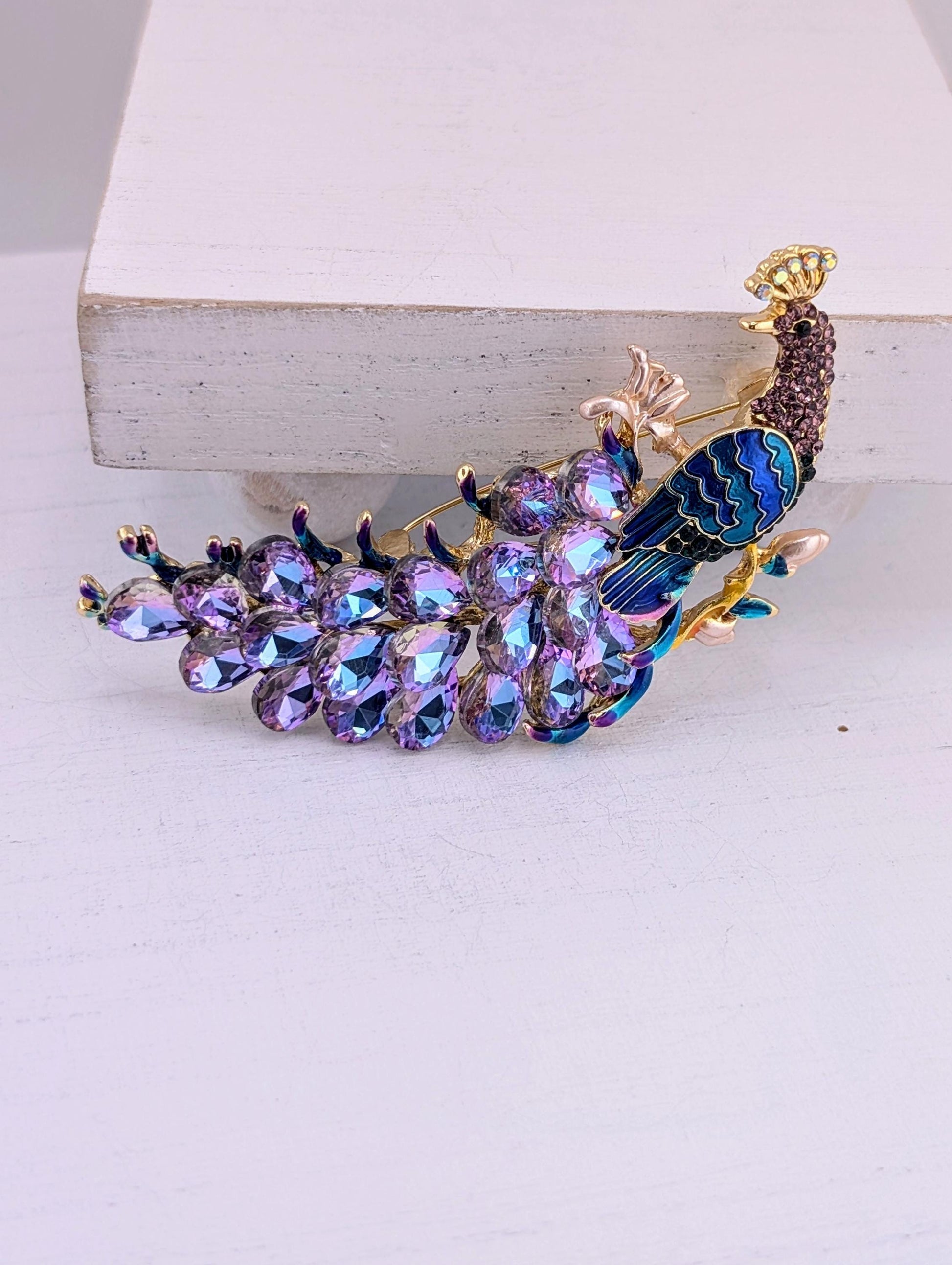 A decorative peacock brooch with a vibrant purple and blue color scheme, adorned with numerous sparkling gemstones.