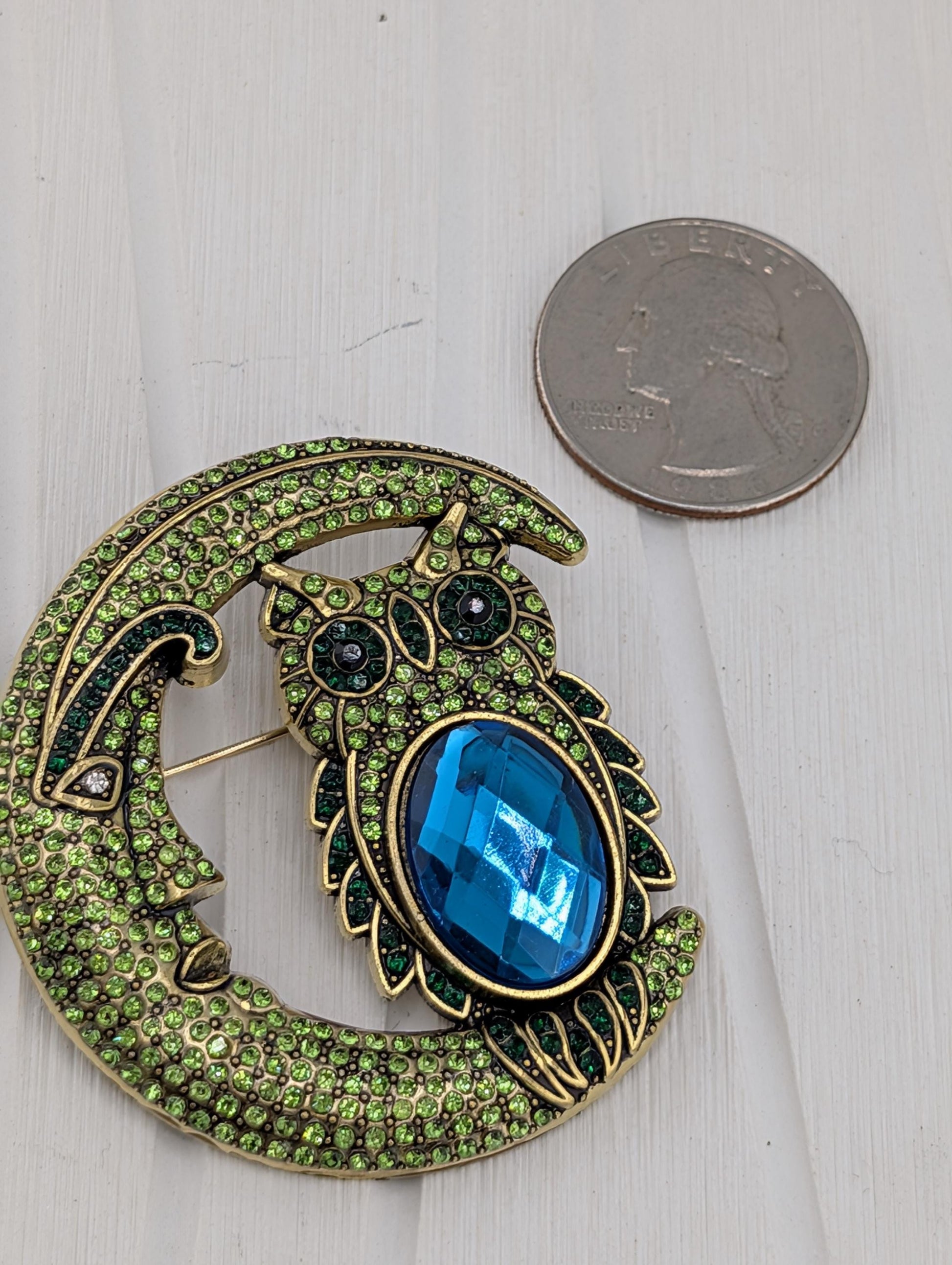 A decorative brooch in the shape of a crescent moon, adorned with green gemstones and a large blue gemstone, placed next to a United States quarter.