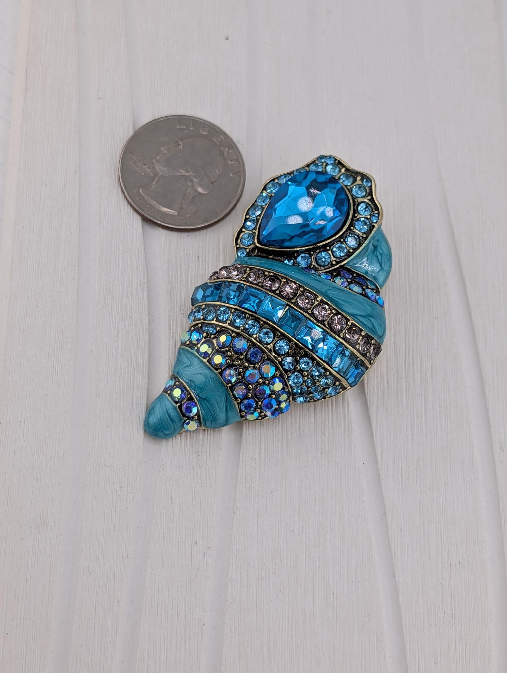 A decorative blue and silver brooch with a heart-shaped gemstone is placed next to a US quarter on a white wooden surface.
