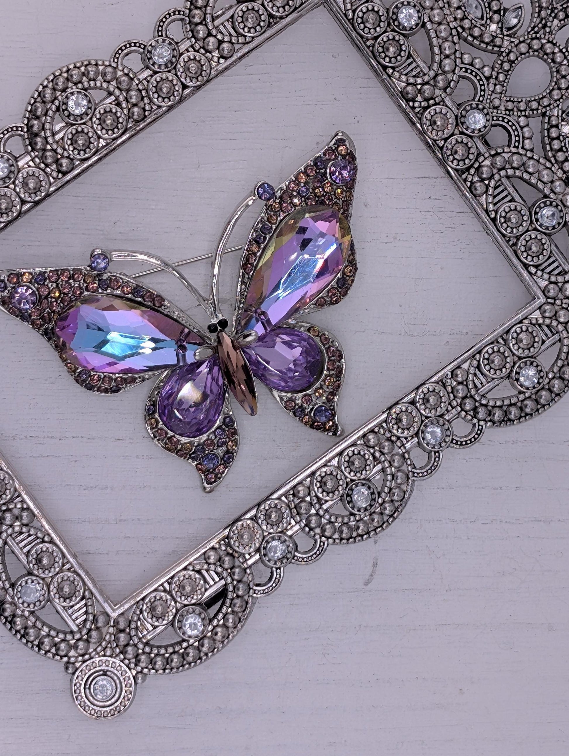 A decorative butterfly brooch with purple and blue gemstones is displayed against a white background, framed by an ornate silver frame.