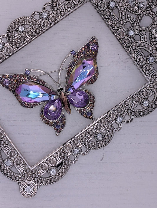 A decorative butterfly brooch with purple and blue gemstones is displayed against a white background, framed by an ornate silver frame.