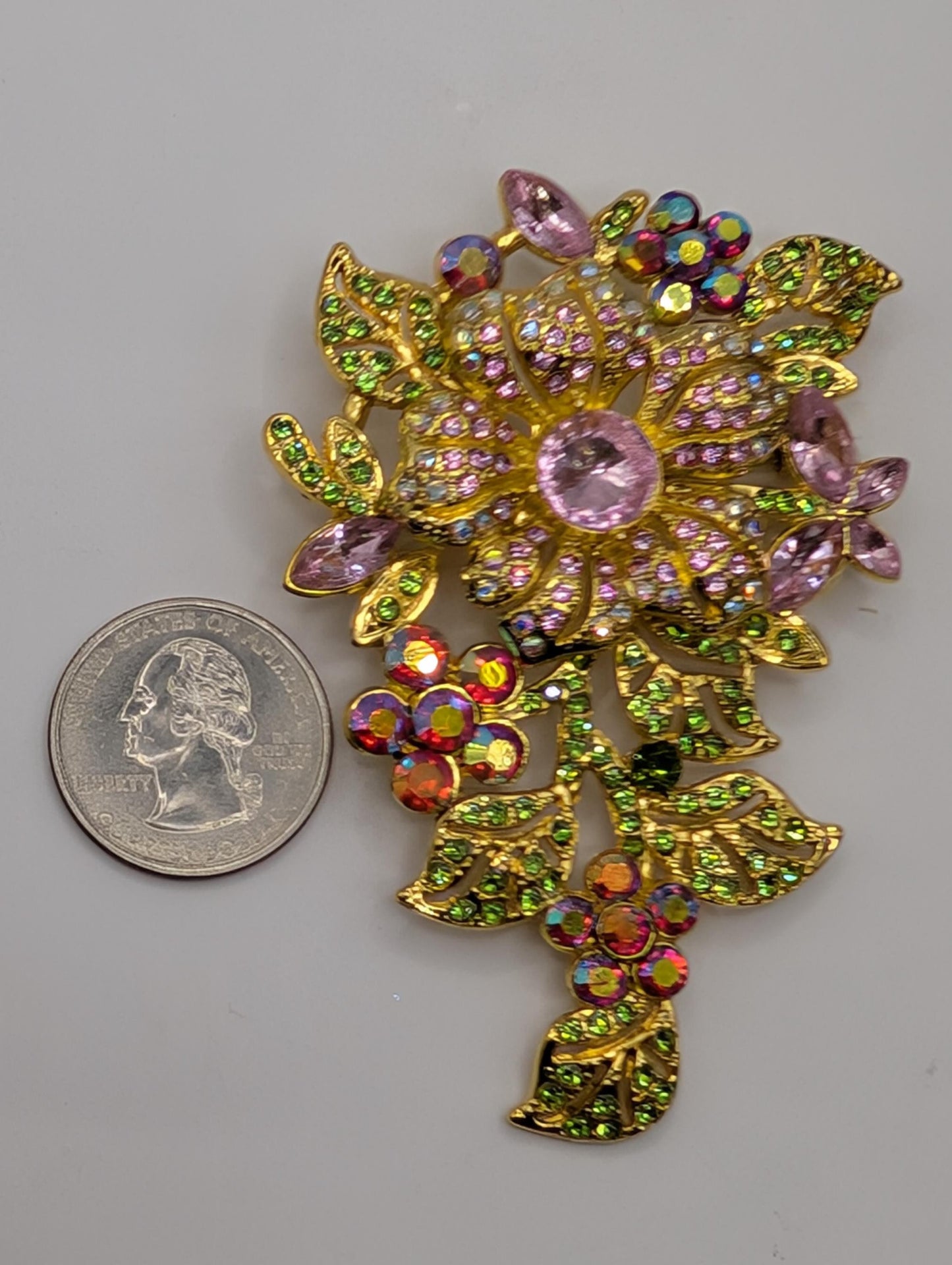 A colorful, ornate brooch adorned with gemstones and leaves is placed next to a United States quarter coin.