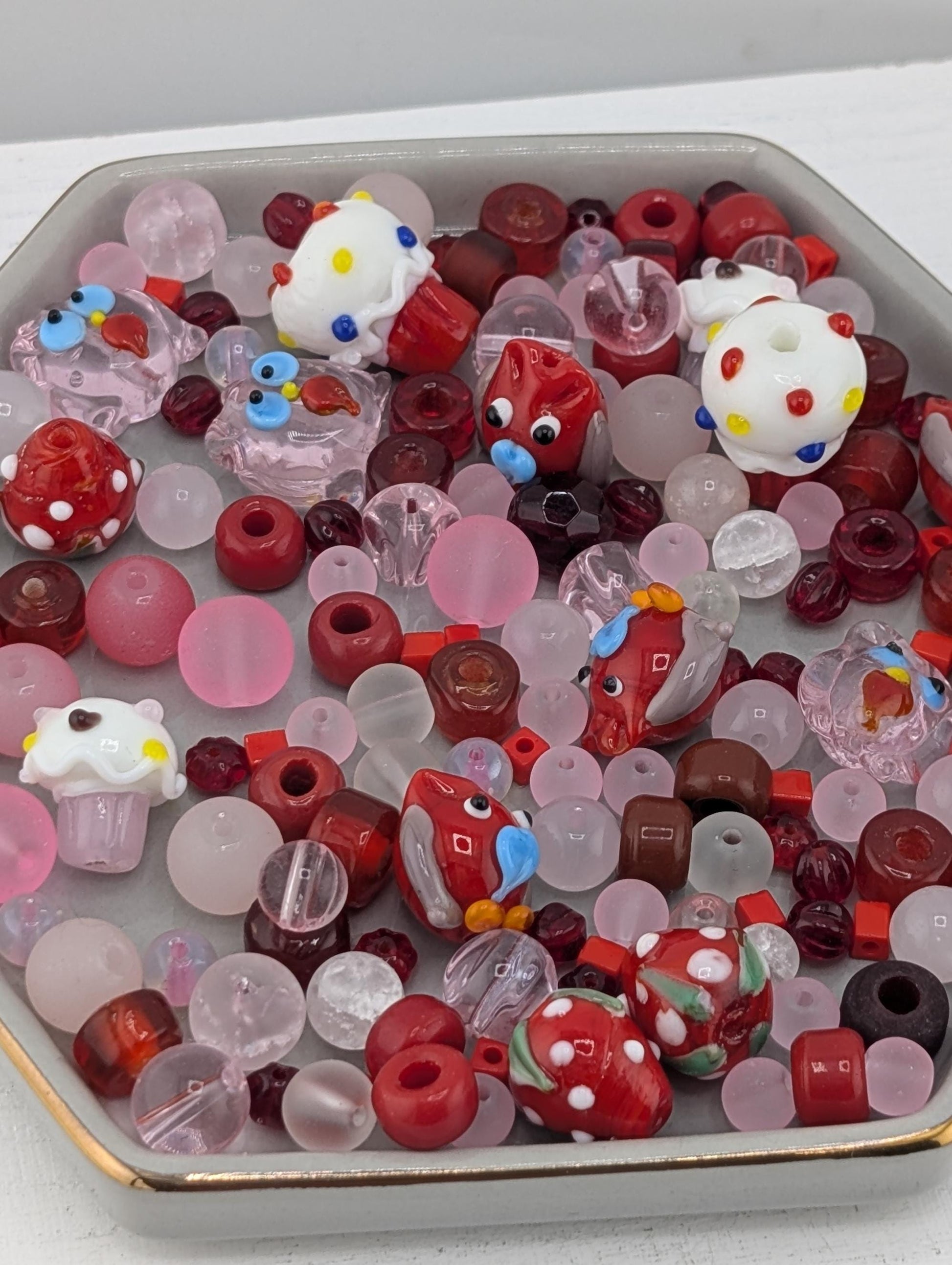 A collection of colorful glass beads, including red, white, and pink beads, as well as some with polka dots and other playful designs, arranged in a container.