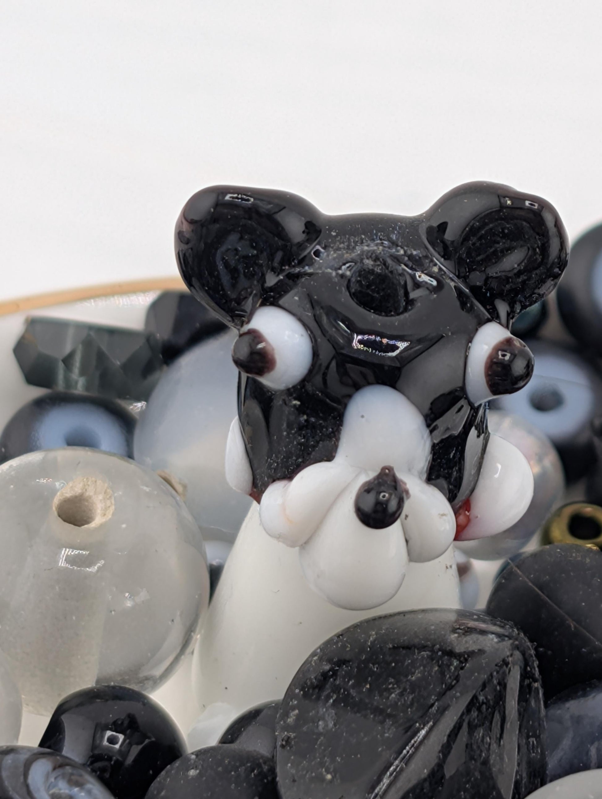 A close-up image of a black and white glass figurine, resembling a dog or a bear, sitting among various glass beads and stones.