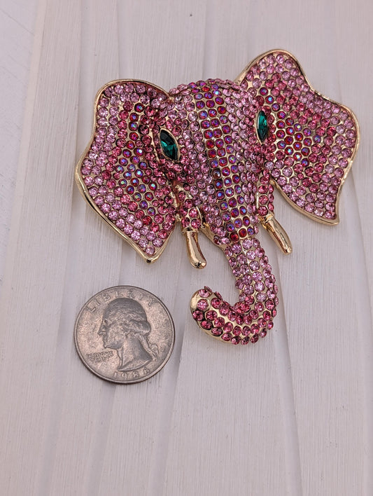 A large, ornate brooch in the shape of an elephant head, adorned with pink and purple gemstones, is displayed next to a United States quarter coin.
