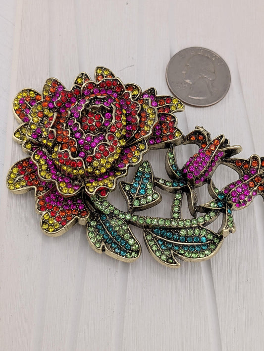 A colorful, sparkling brooch with a large, vibrant flower design and various gemstones.