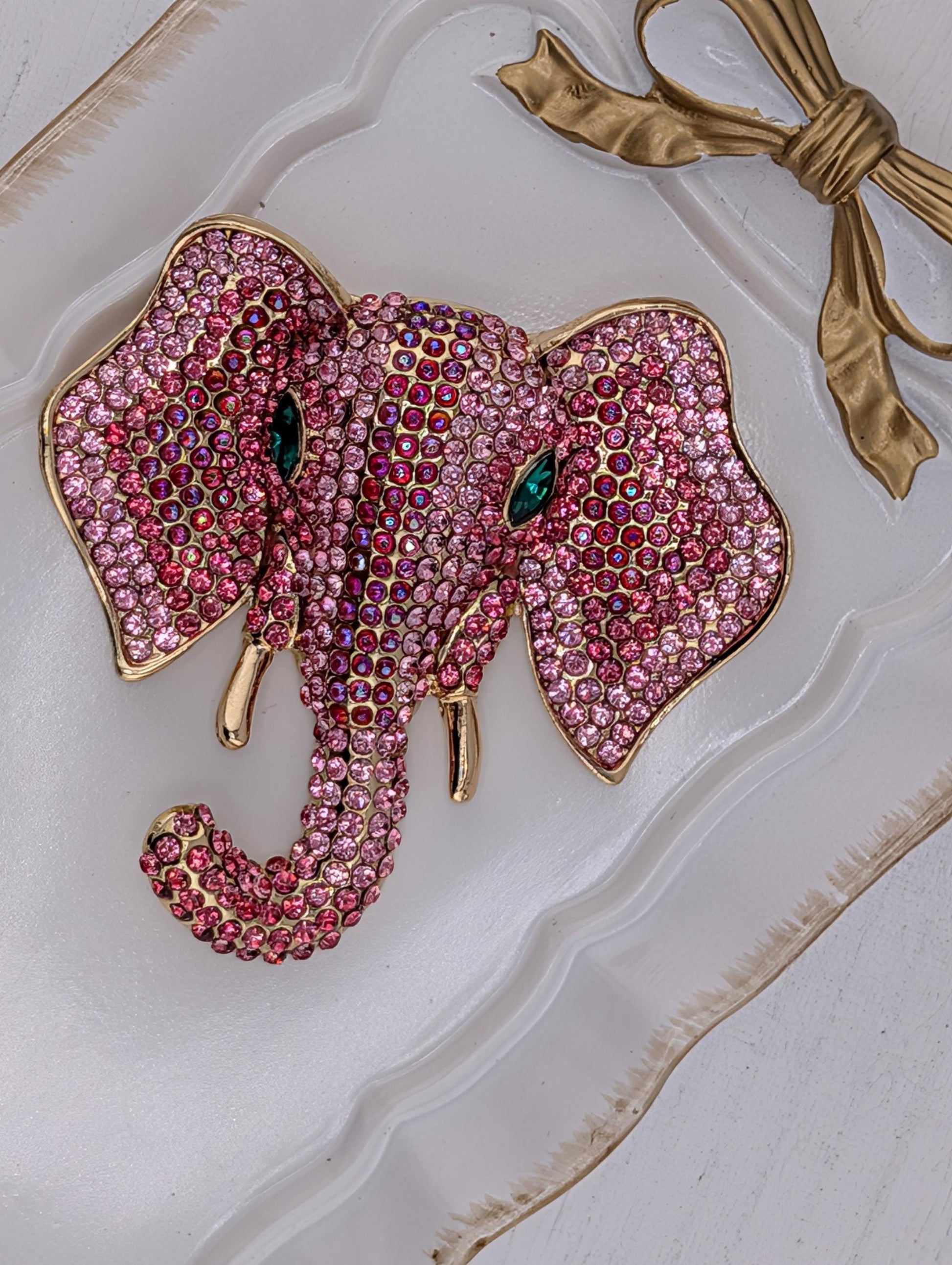 A pink elephant brooch adorned with sparkling rhinestones and a gold bow is displayed on a white surface.