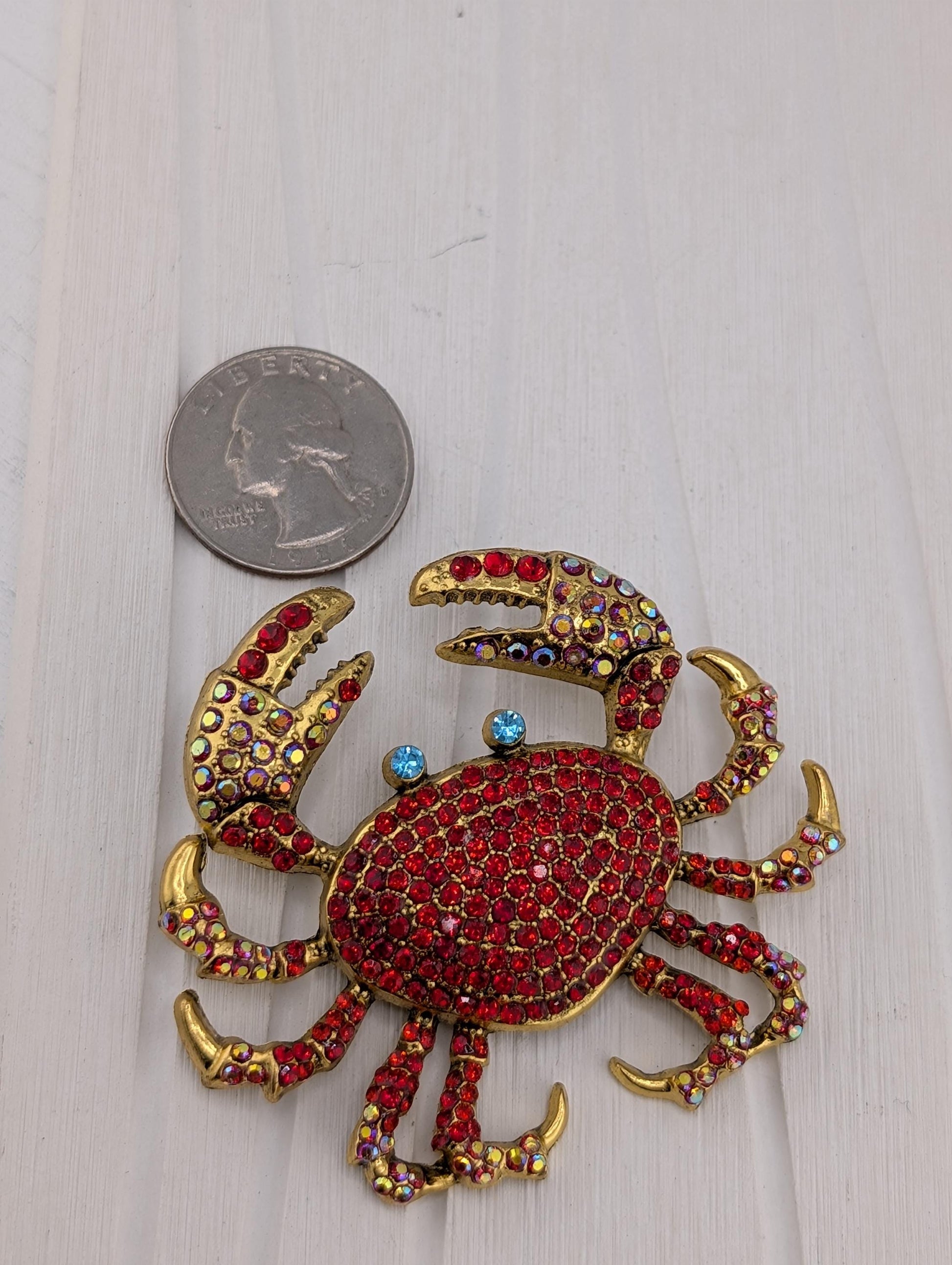 A shiny, ornate crab brooch with red and gold jewels is placed next to a United States quarter coin on a white surface.