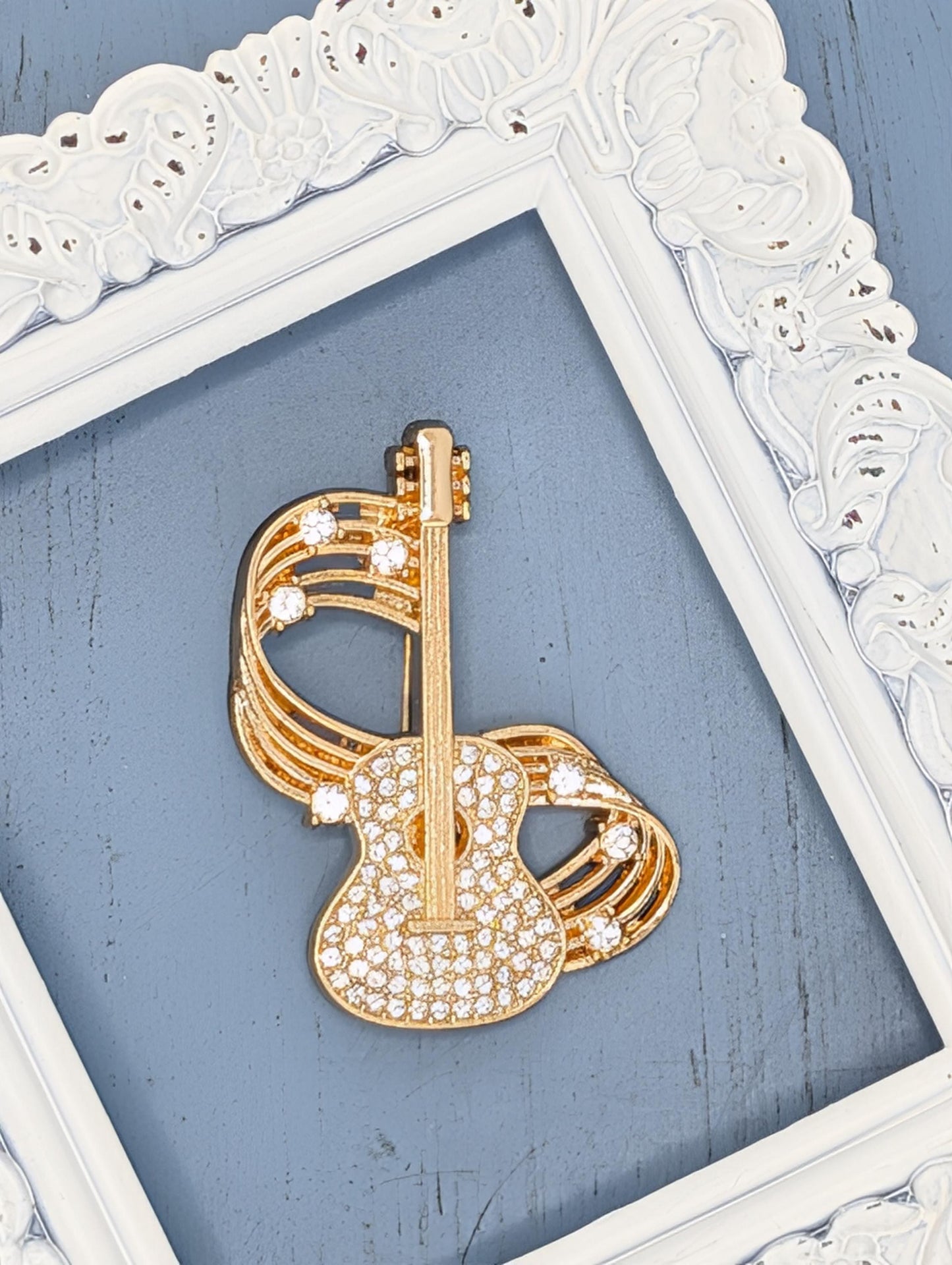 A gold-colored guitar brooch with rhinestones is displayed against a blue background, framed by a white ornate frame.