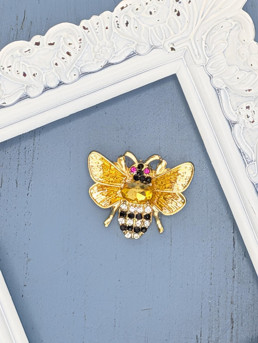 A decorative gold-colored bee brooch with a black and white striped pattern and a yellow center is displayed against a blue background.