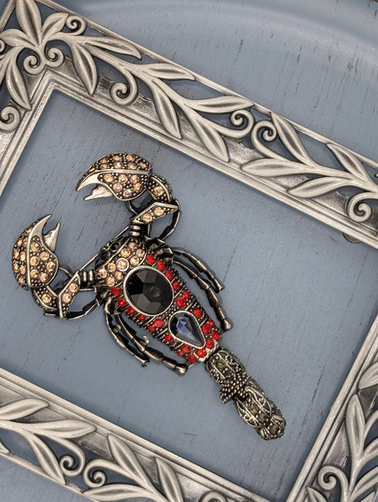 A decorative metal frame with a framed metal sculpture of a scorpion, adorned with gemstones and intricate designs.