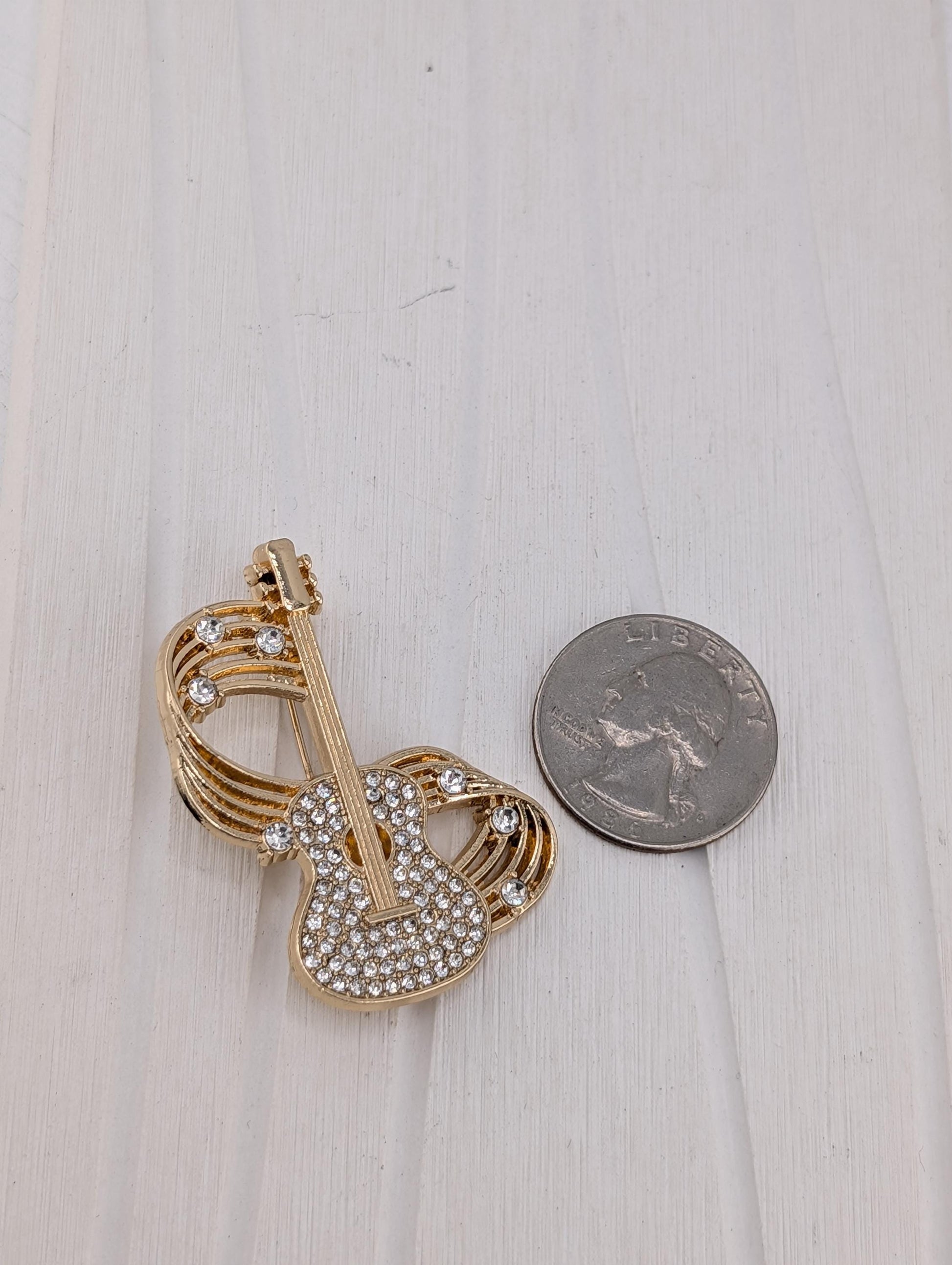 A gold-colored brooch with a guitar-shaped design and sparkling rhinestones is placed next to a silver coin on a white wooden surface.