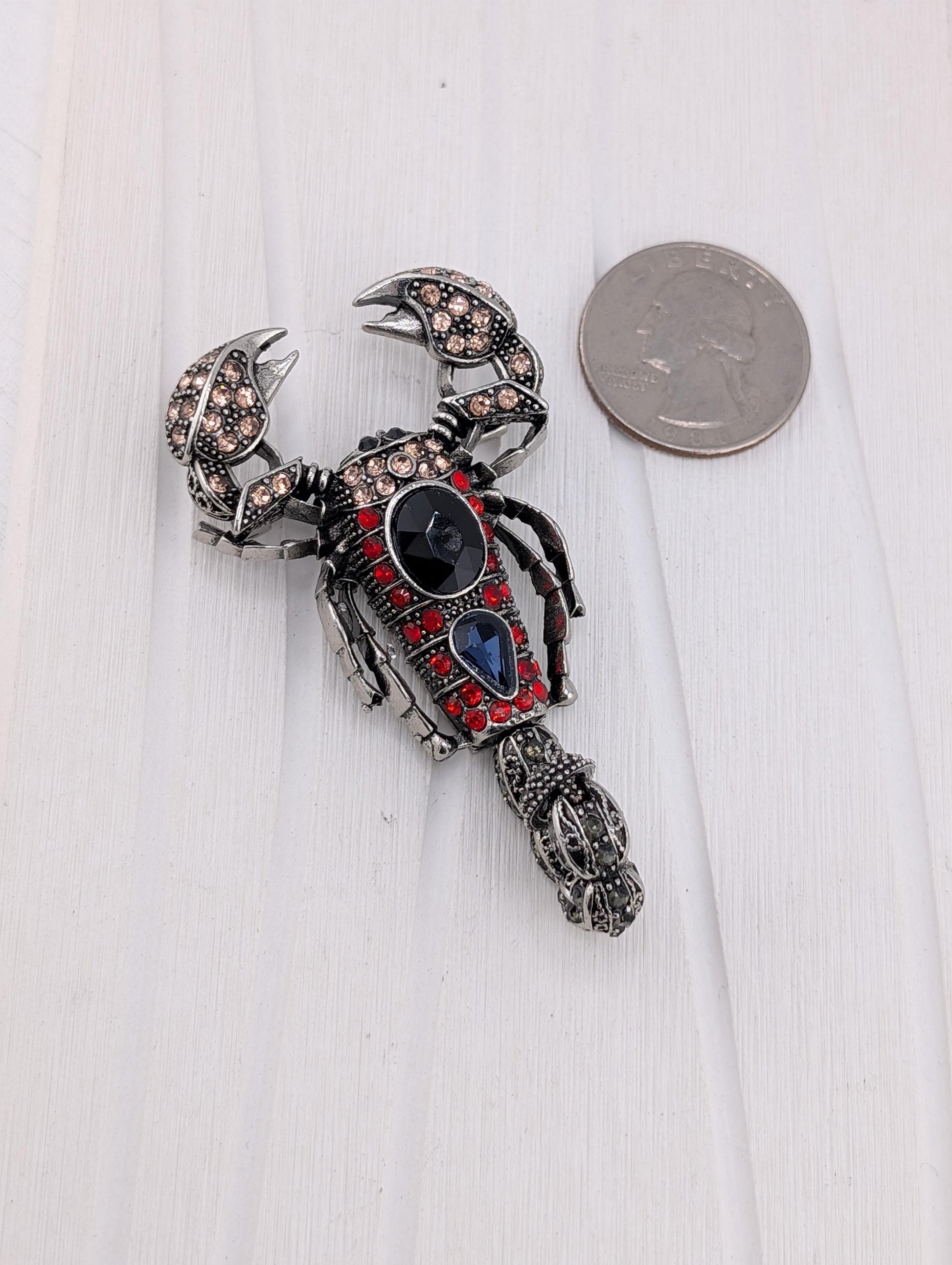 A decorative metal lobster brooch with a red and black color scheme, adorned with gemstones, is placed next to a US quarter on a white surface.