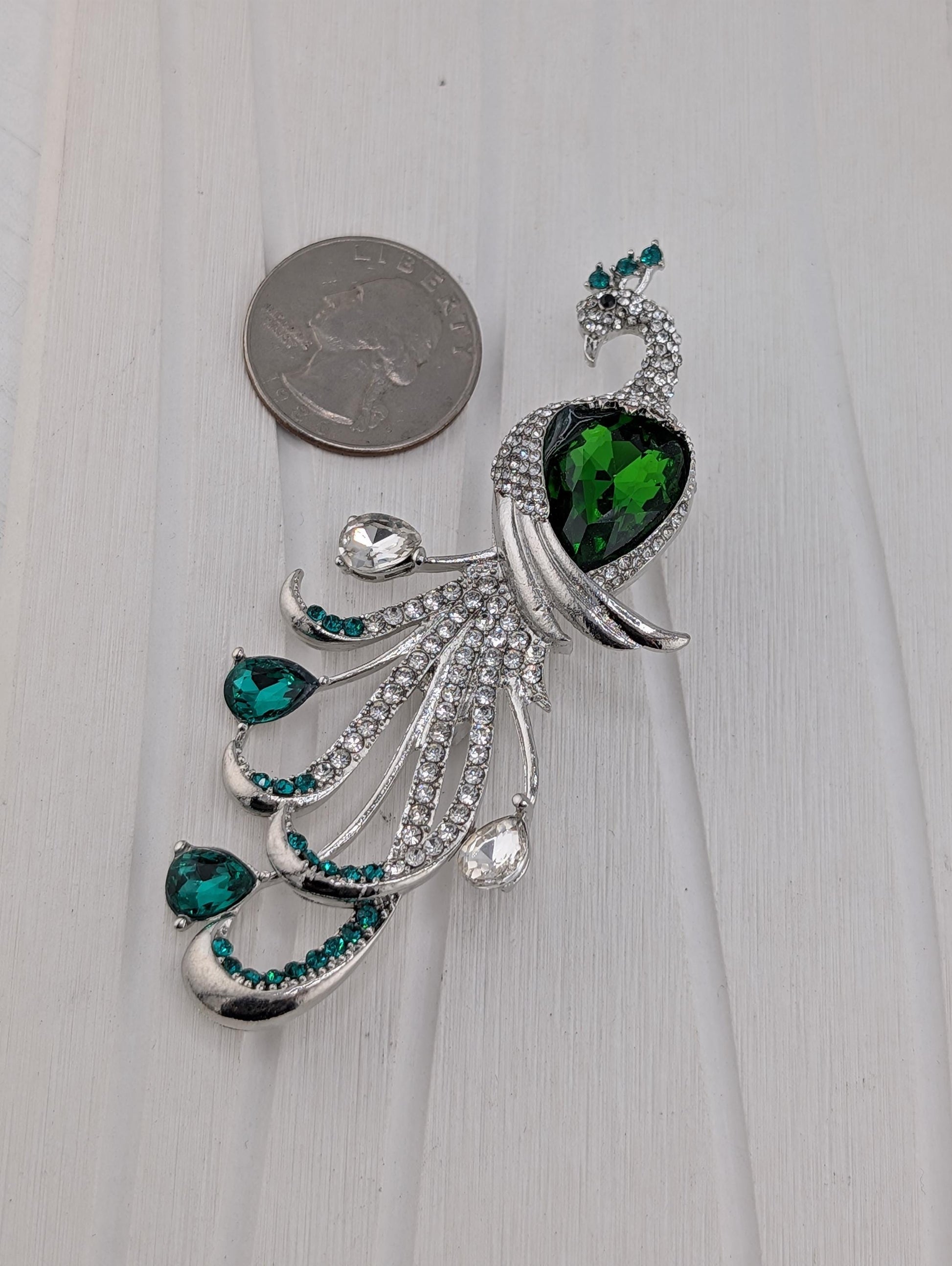 A silver brooch with a peacock design, featuring a large green gemstone and smaller green gemstones, is placed next to a coin on a white surface.
