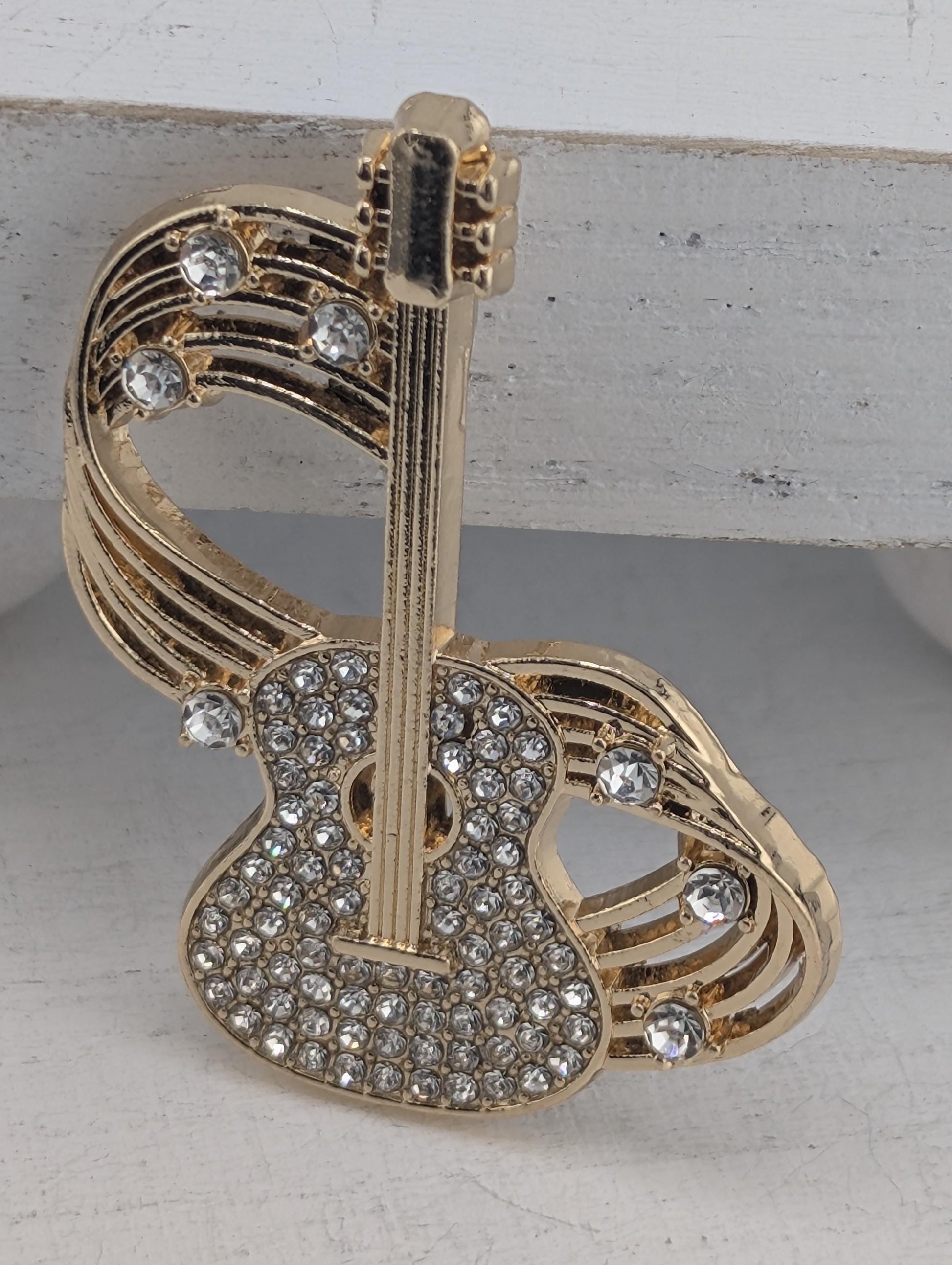 A gold-colored pin or brooch with a guitar-shaped design and sparkling rhinestones or crystals on it.